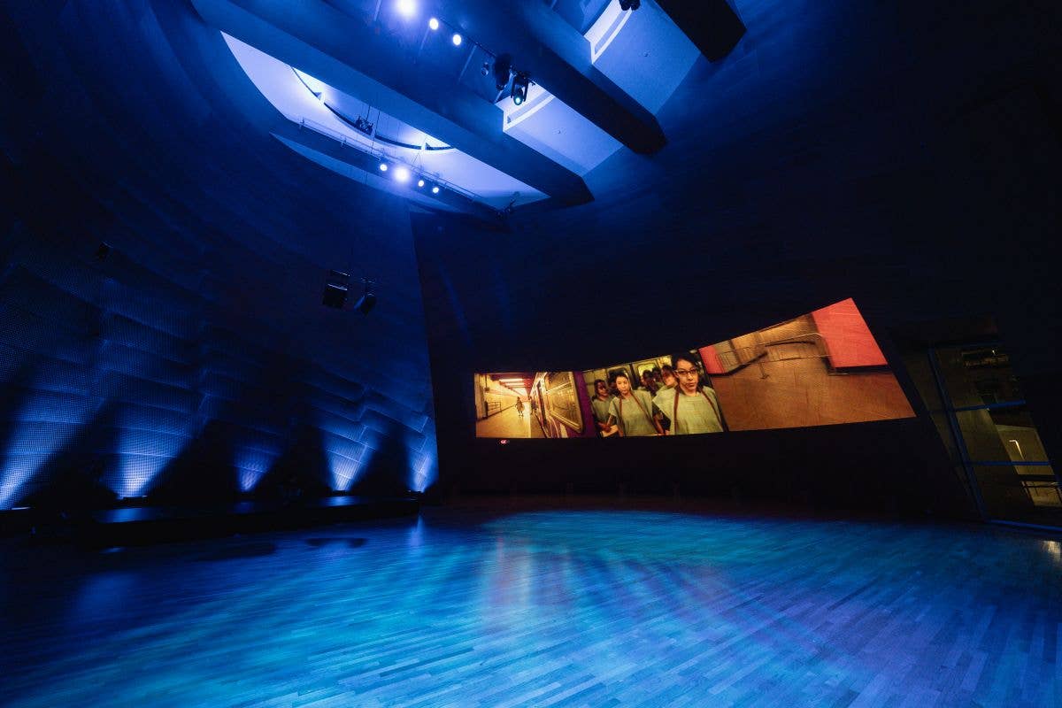 Estudio de teatro a oscuras con iluminación azul, escenario vacío y una pantalla en la pared en la que se ven personas y paneles de colores.