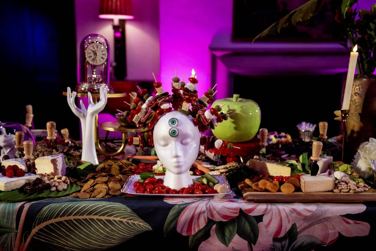 Escena festiva y surrealista en una mesa, con una escultura de una cabeza decorada, velas, frutas y un reloj iluminado en morado al fondo