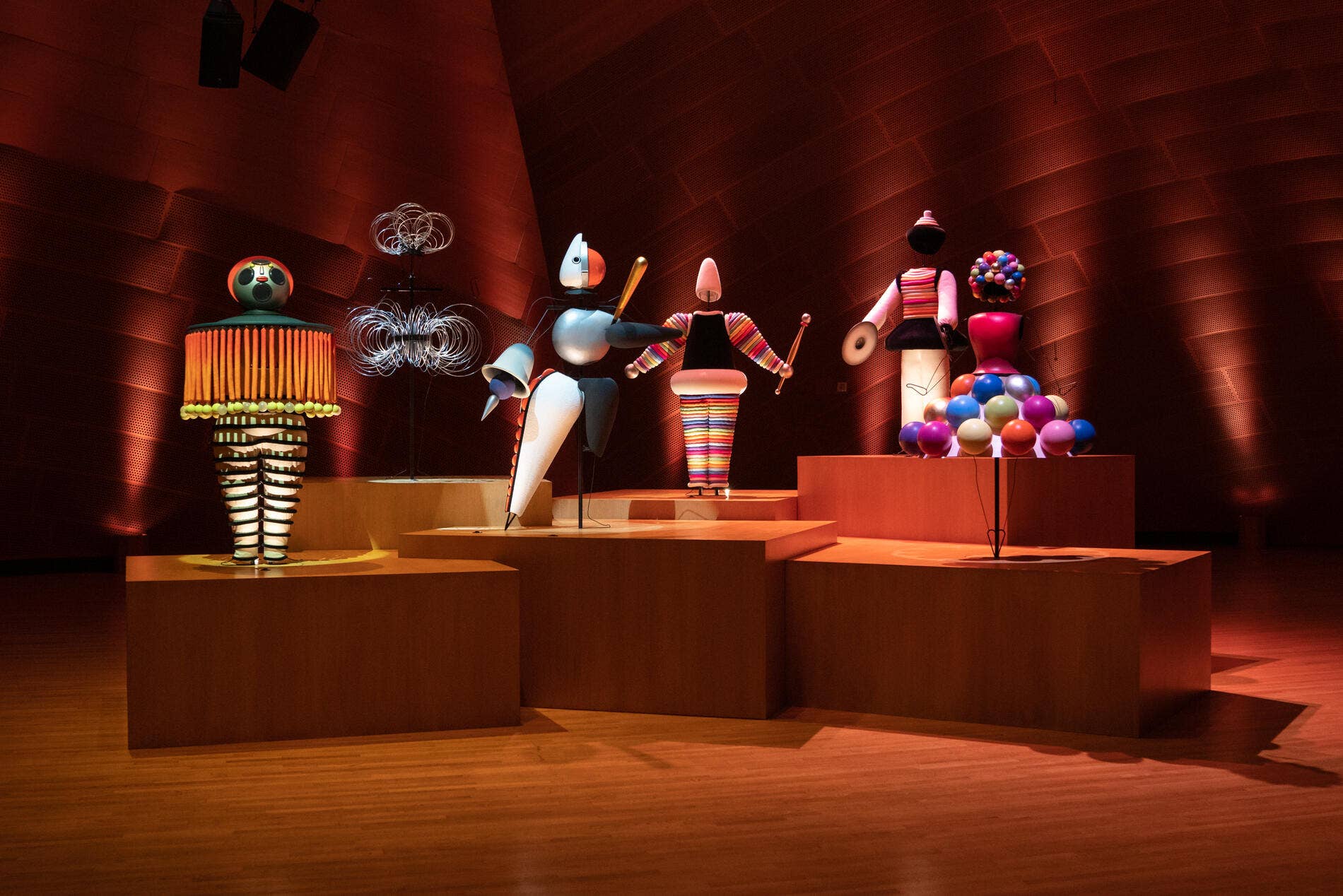 Esculturas de robots de colores sobre un pedestal de galería, bajo una cálida iluminación roja, en una sala de exposiciones.