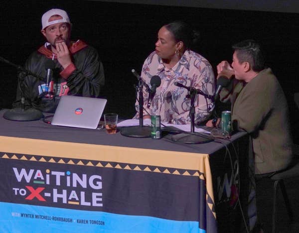 Tres personas están sentadas en una mesa de podcast con micrófonos y un ordenador portátil; en la pancarta se lee «Waiting to X-Hale».