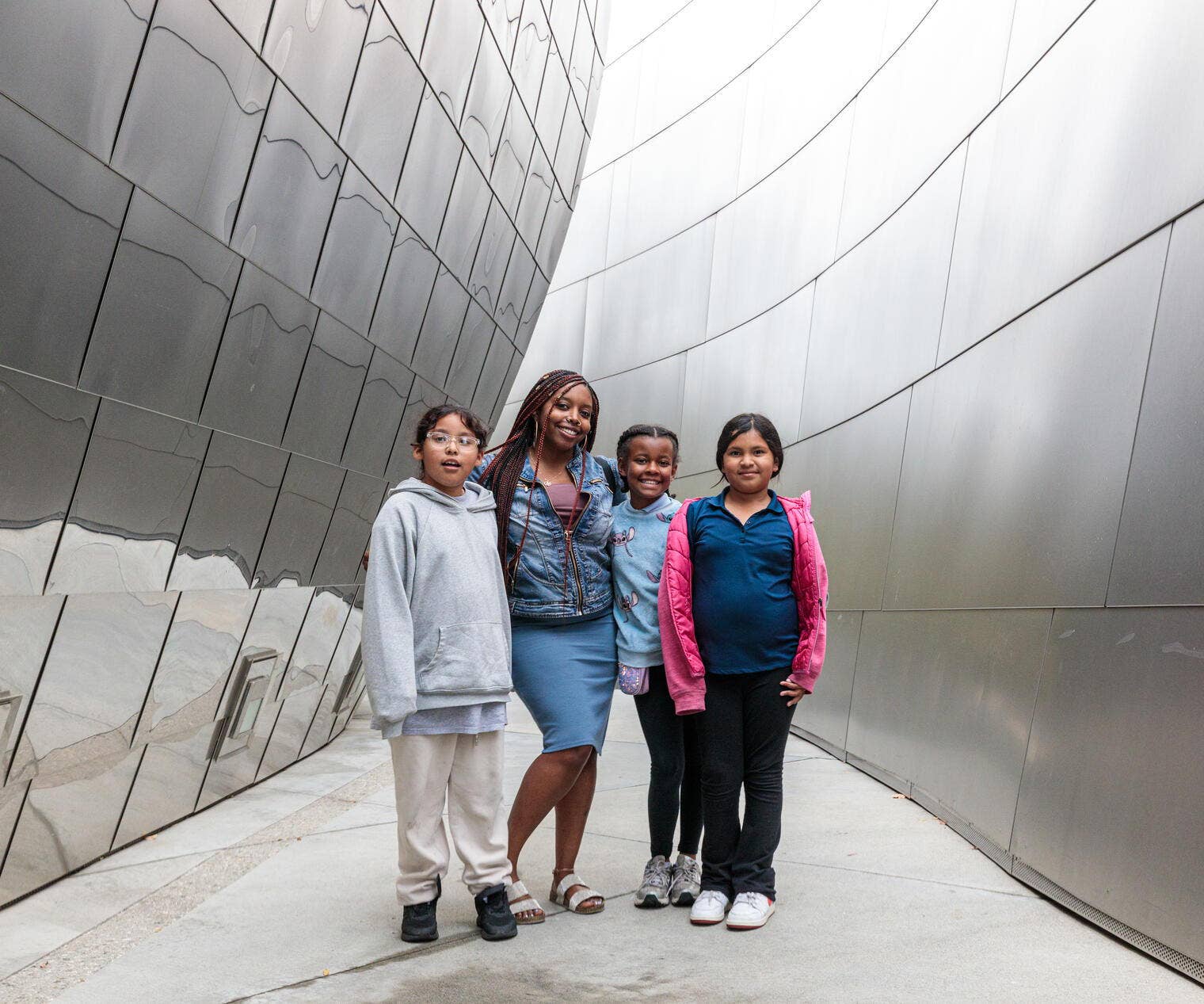 Four children pose together against curved gray stone walls, smiling in casual hoodies and denim.