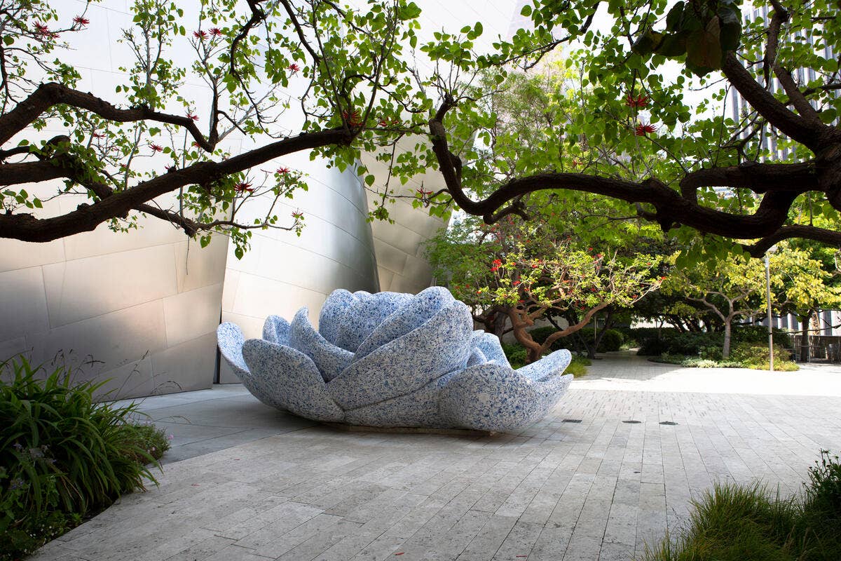 Un asiento escultural de color azul, con aspecto de piedra, en forma de espiral bajo unas ramas frondosas, junto a un muro blanco curvo en el patio ajardinado