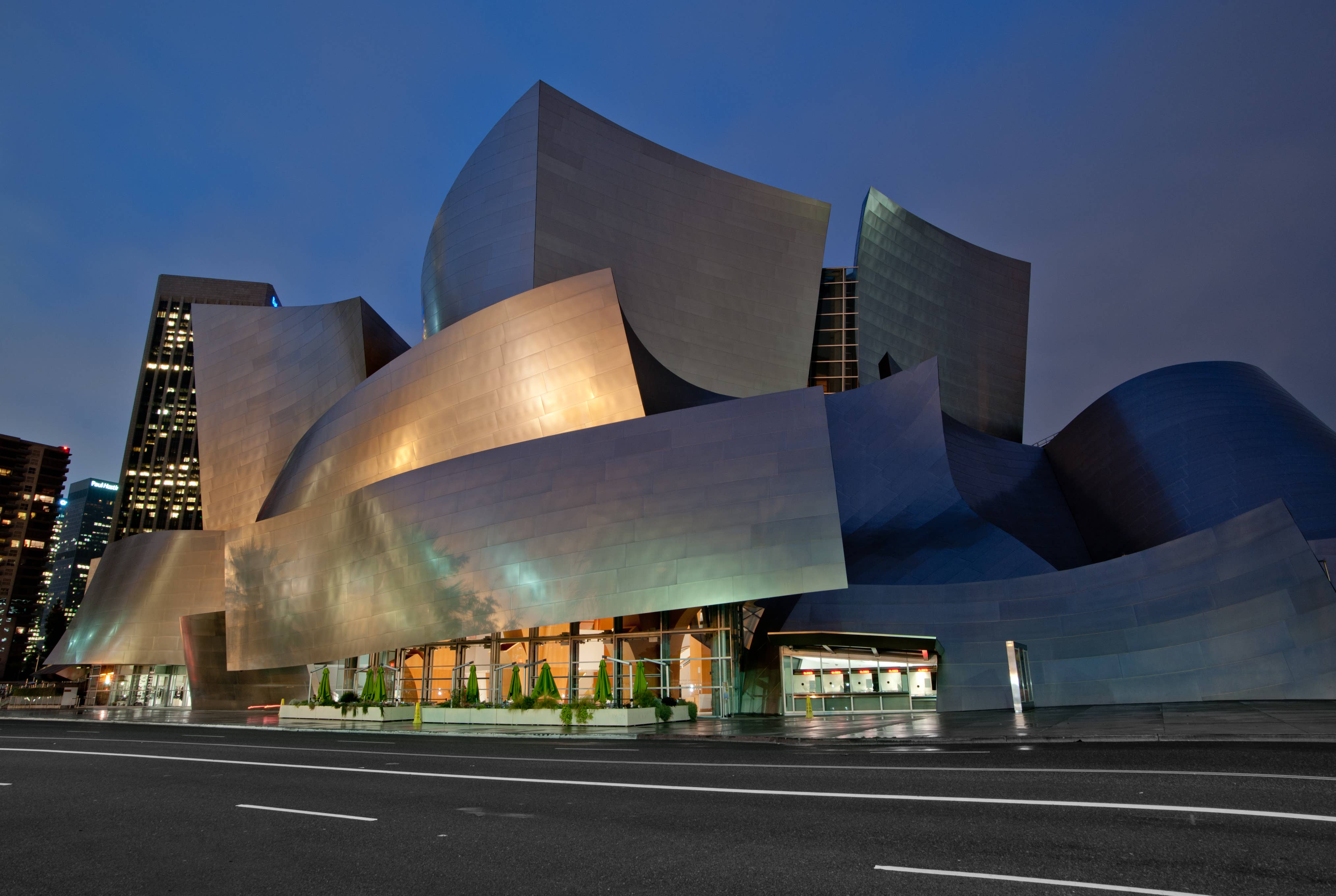 Modern museum facade with curved metal panels and glowing windows at dusk, with city buildings nearby