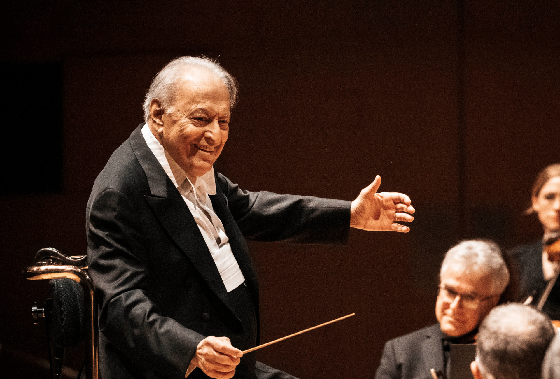Zubin Mehta, vestido con un traje oscuro, gesticula mientras sostiene la batuta  director de orquesta batuta durante una actuación en directo.
