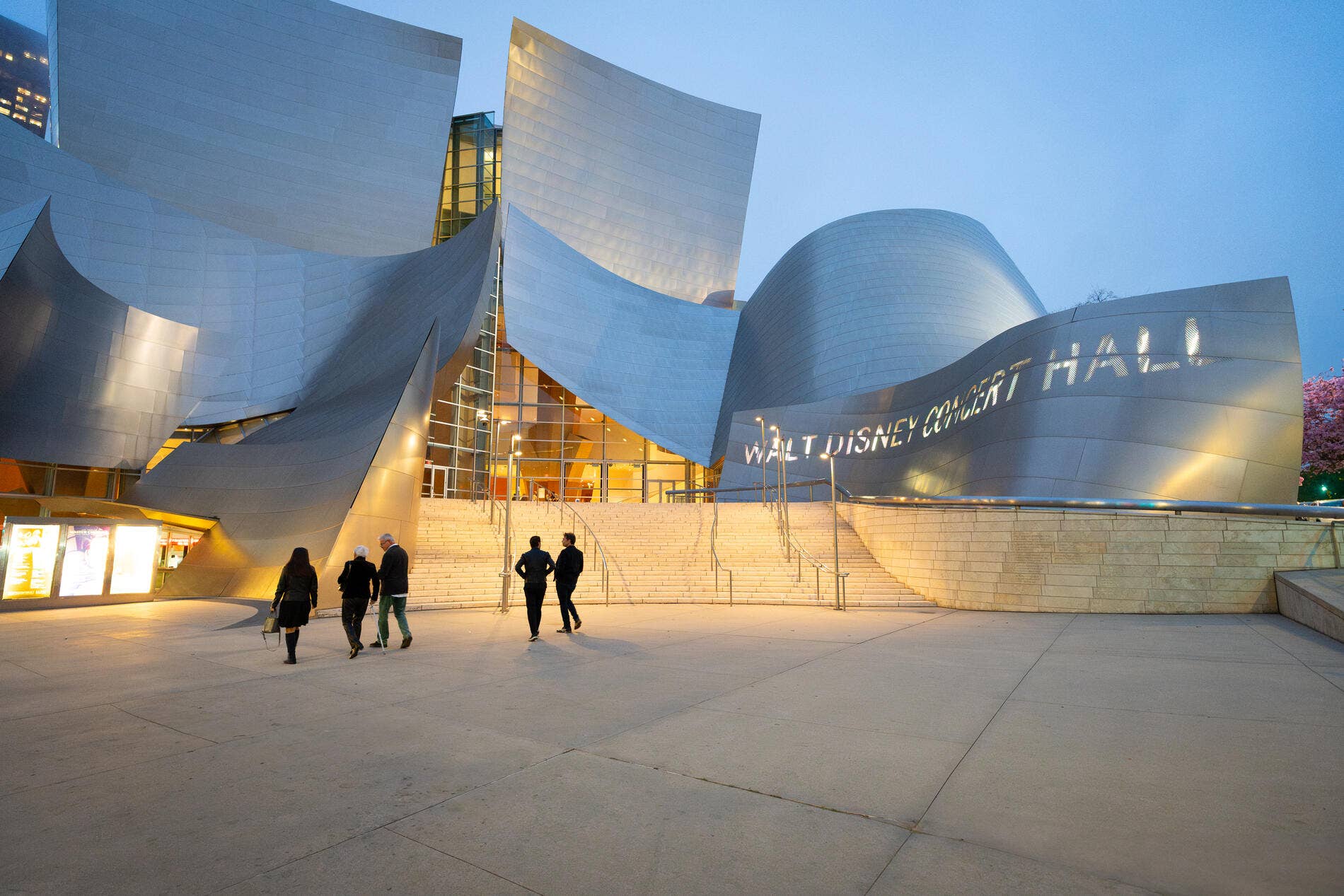 Walt Disney Concert Hall building exterior with curved glass facade, steps, and pedestrians at dusk