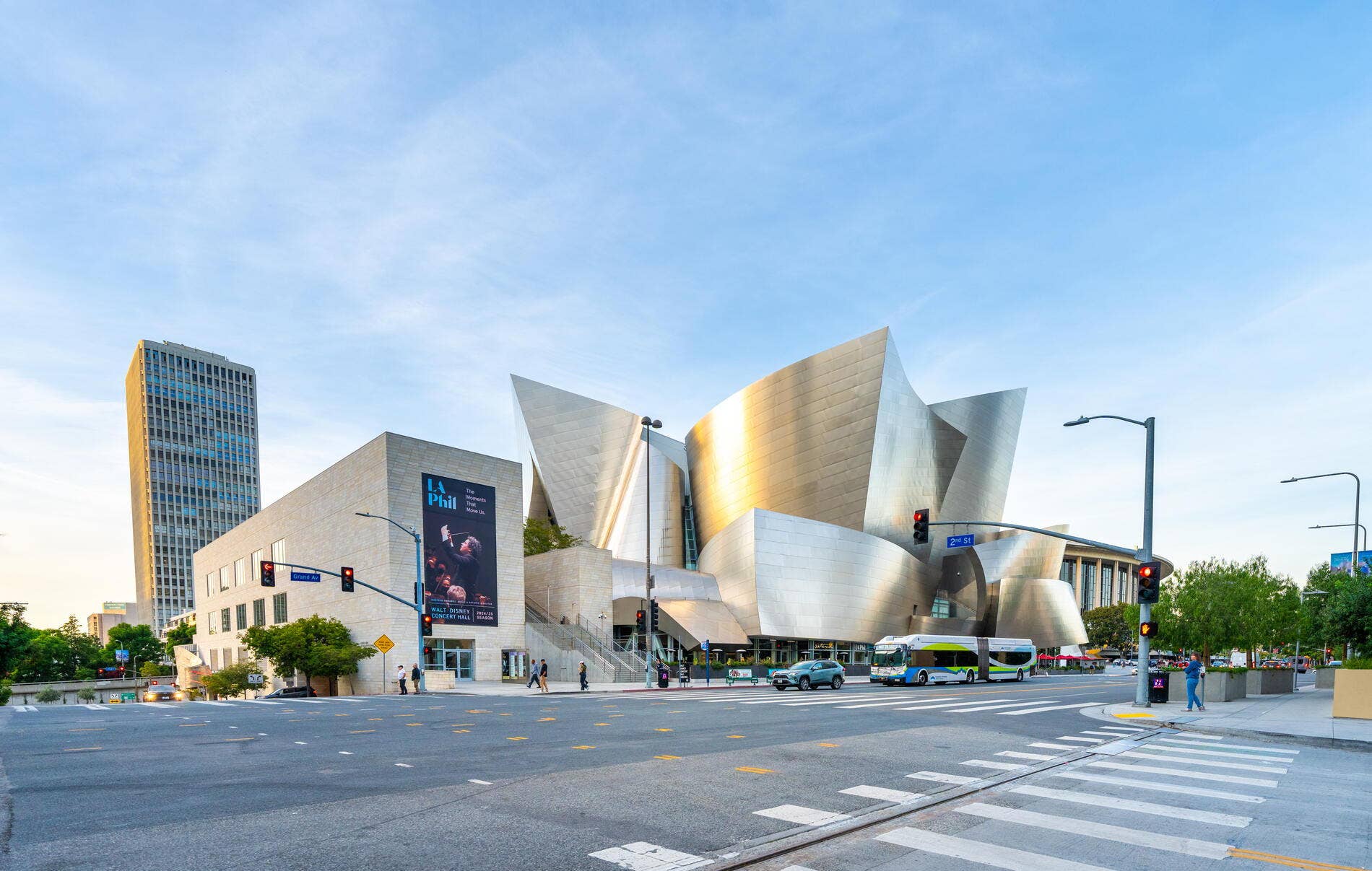 Modern concert hall with sweeping sandstone walls and billboards, viewed from a city intersection under a blue sky