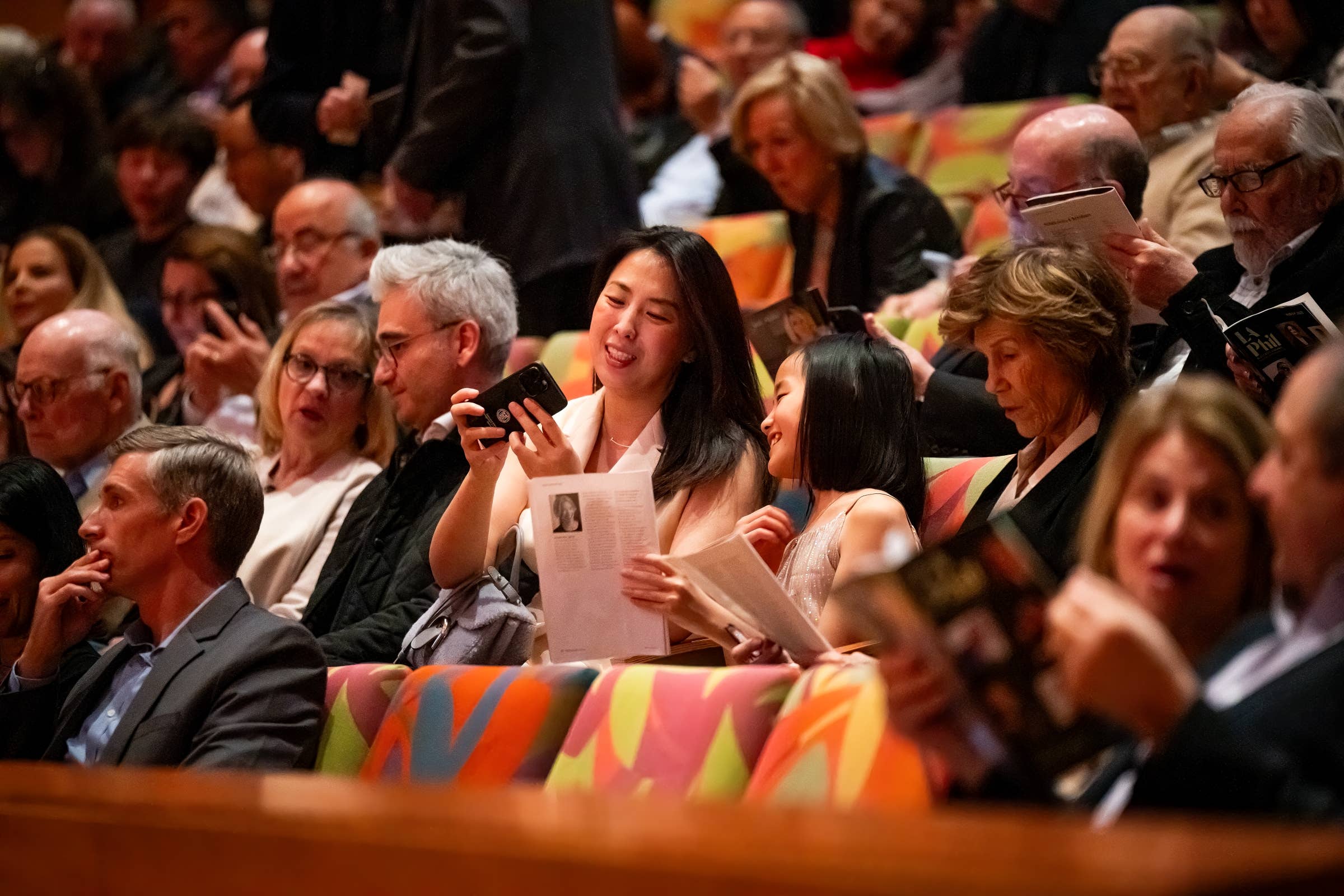 Los asistentes a un auditorio sonríen y aplauden mientras sostienen los programas y utilizan sus teléfonos móviles.