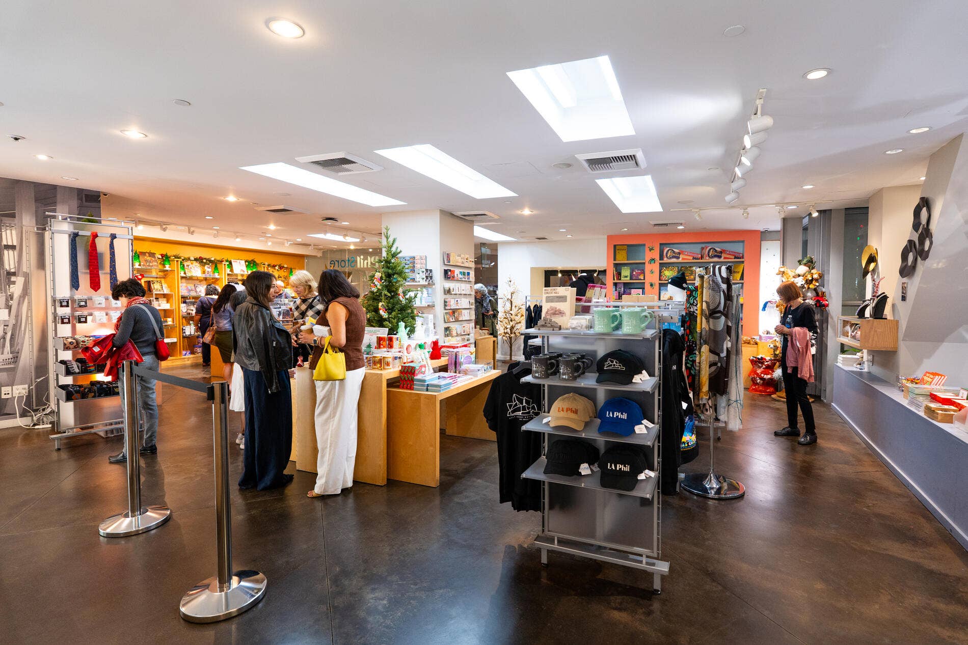 People browsing in a bright retail shop with festive displays, books, mugs, and clothing racks under ceiling lights.