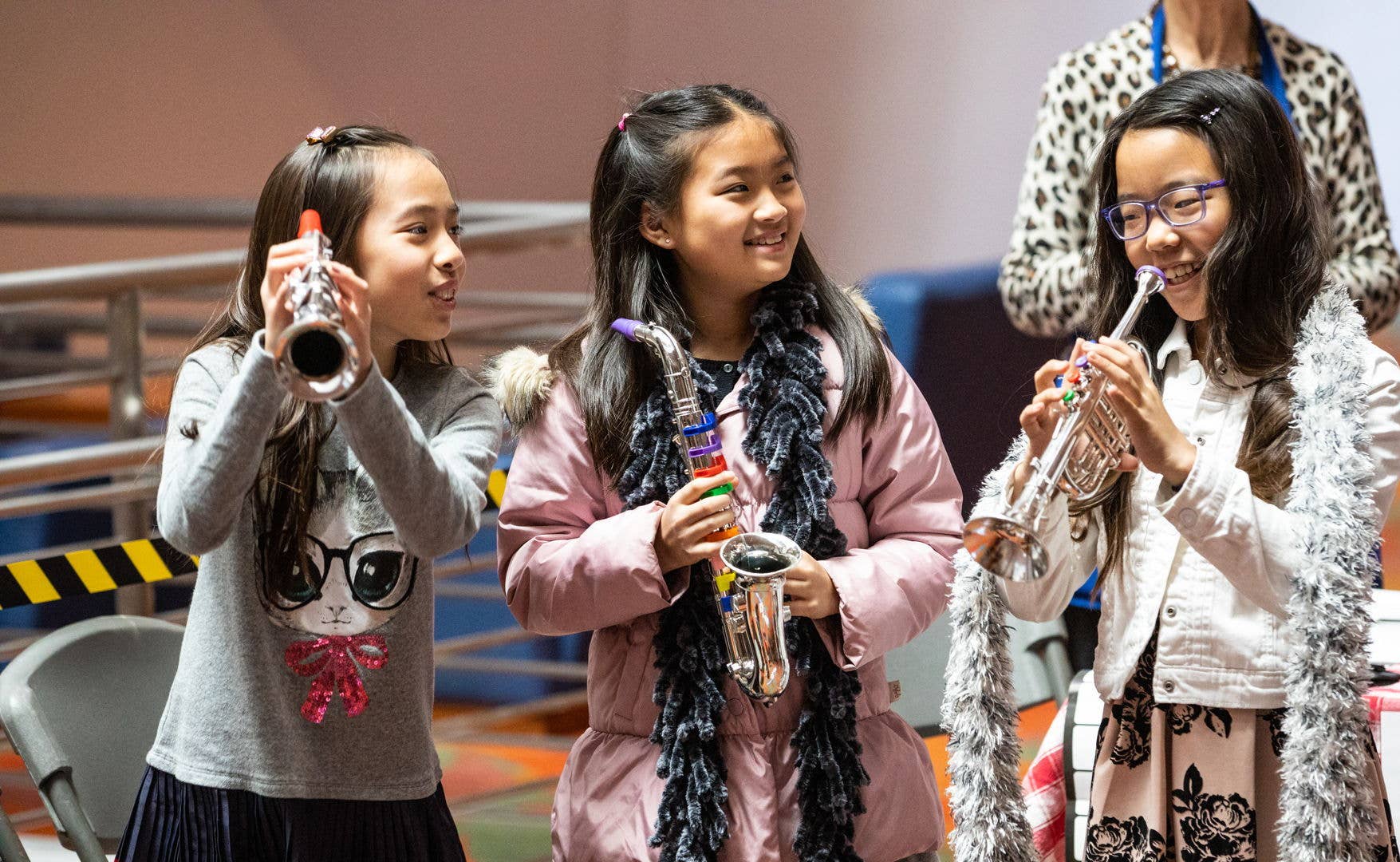Three girls play instruments—one trumpet, one saxophone, and another trumpet—while smiling in a classroom.