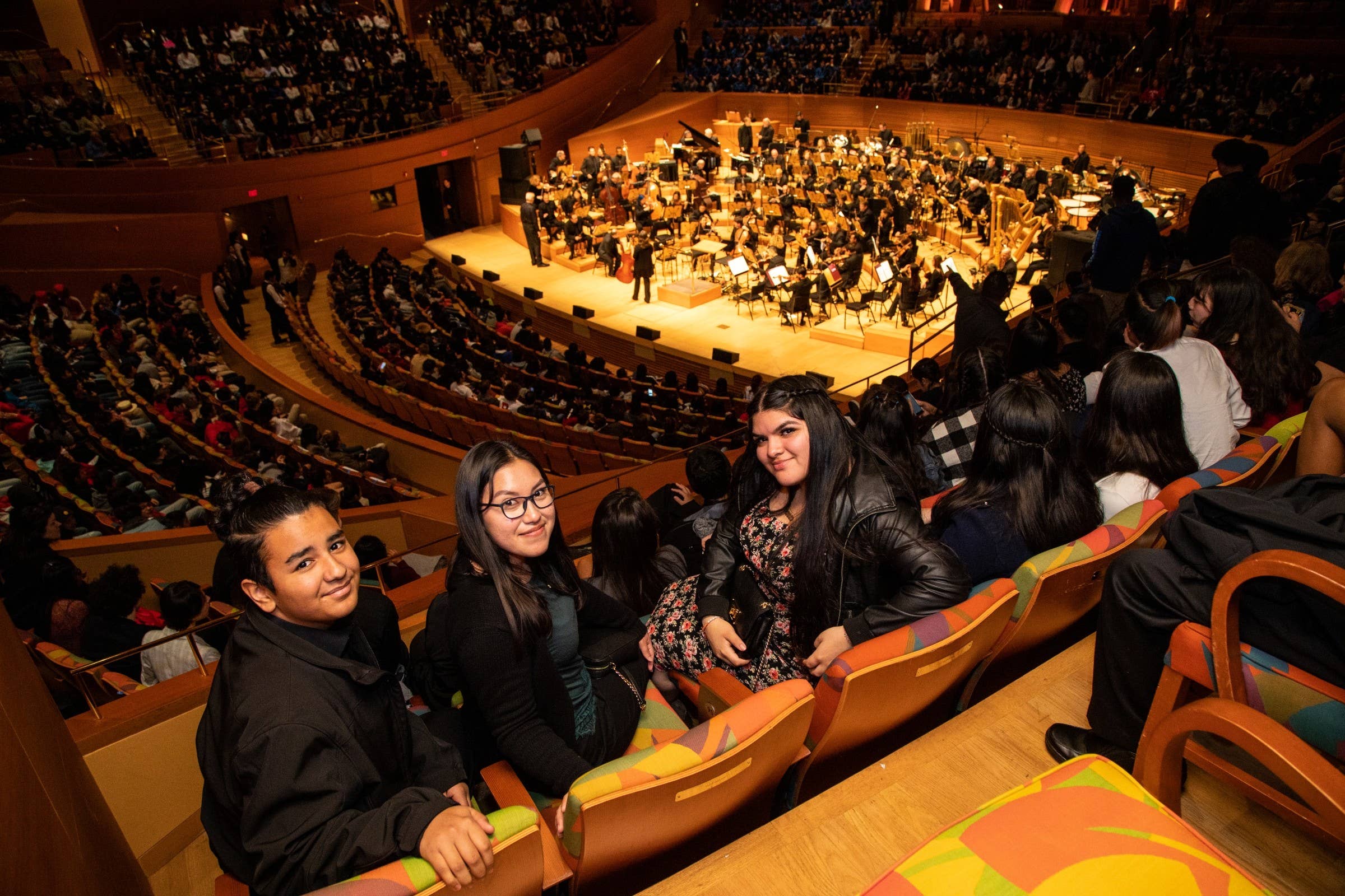 Audience members sit in a theater box while an orchestra performs on stage under warm lights.