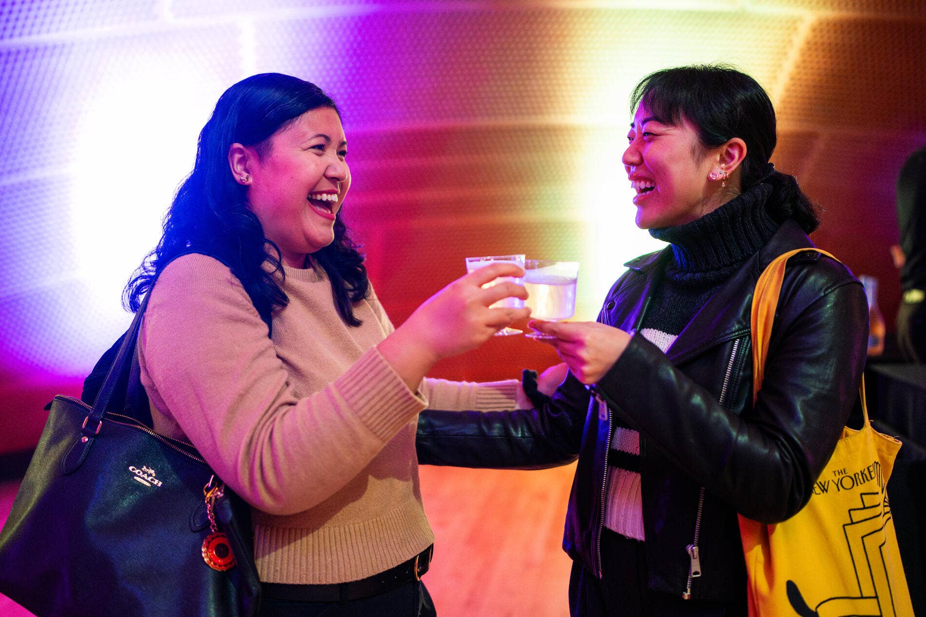 Two women toast with small cups, laughing together at a colorful event with rainbow stage lighting.
