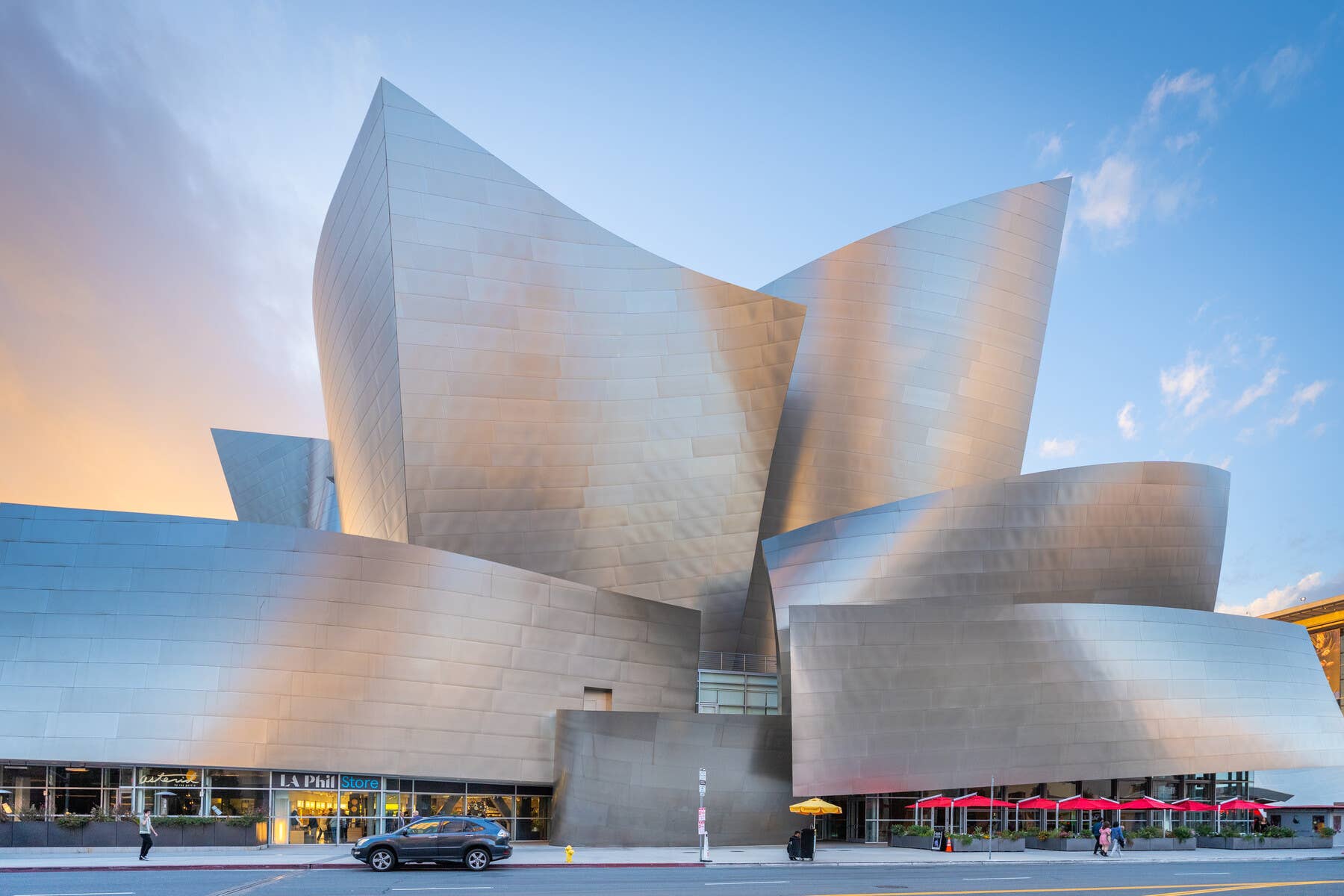 Fachada futurista de una sala de conciertos blanca, con paneles curvos y cristal, al atardecer bajo un cielo azul con nubes