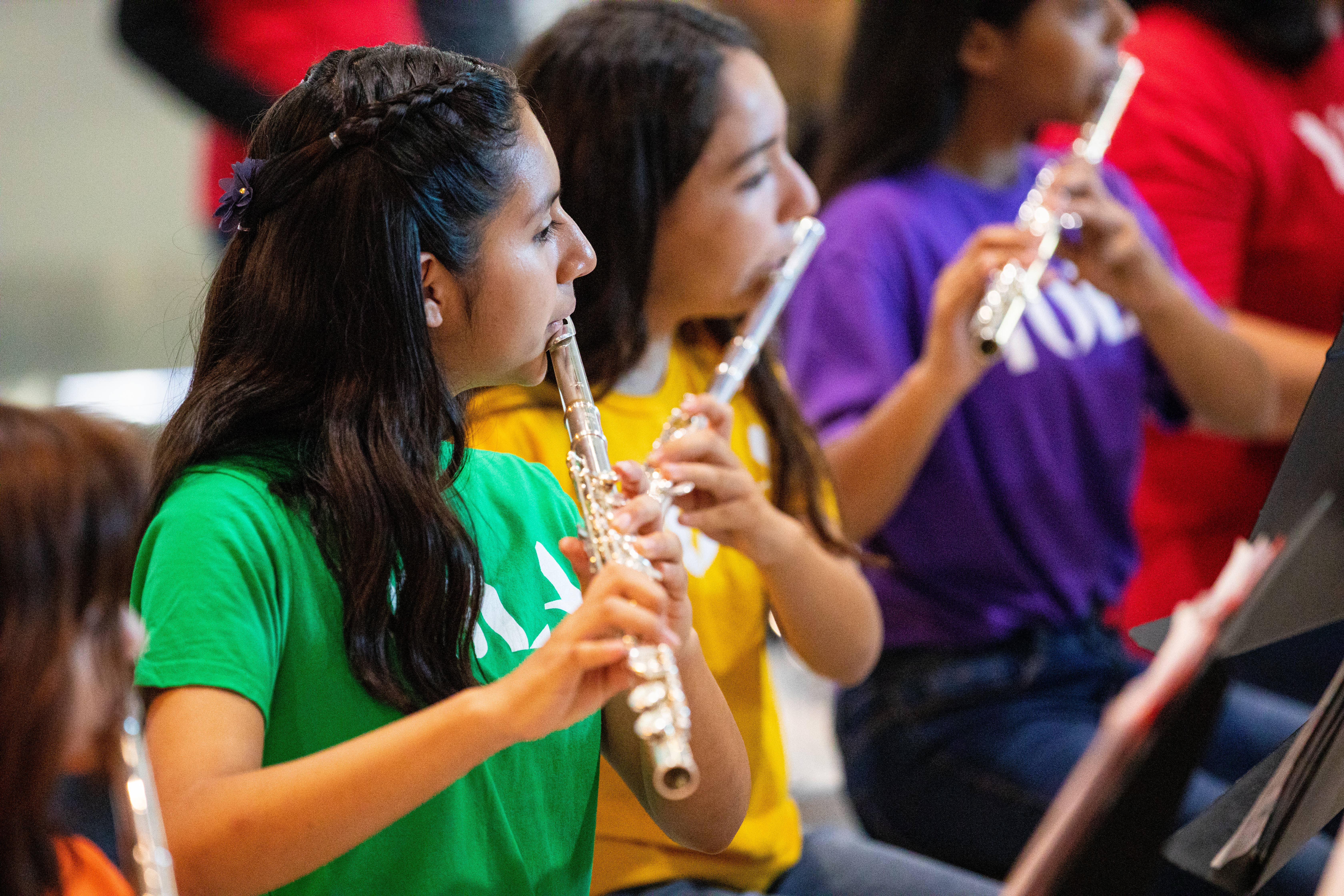 Two women play silver flutes in a colorful band, seen in profile with blurred musicians behind them.