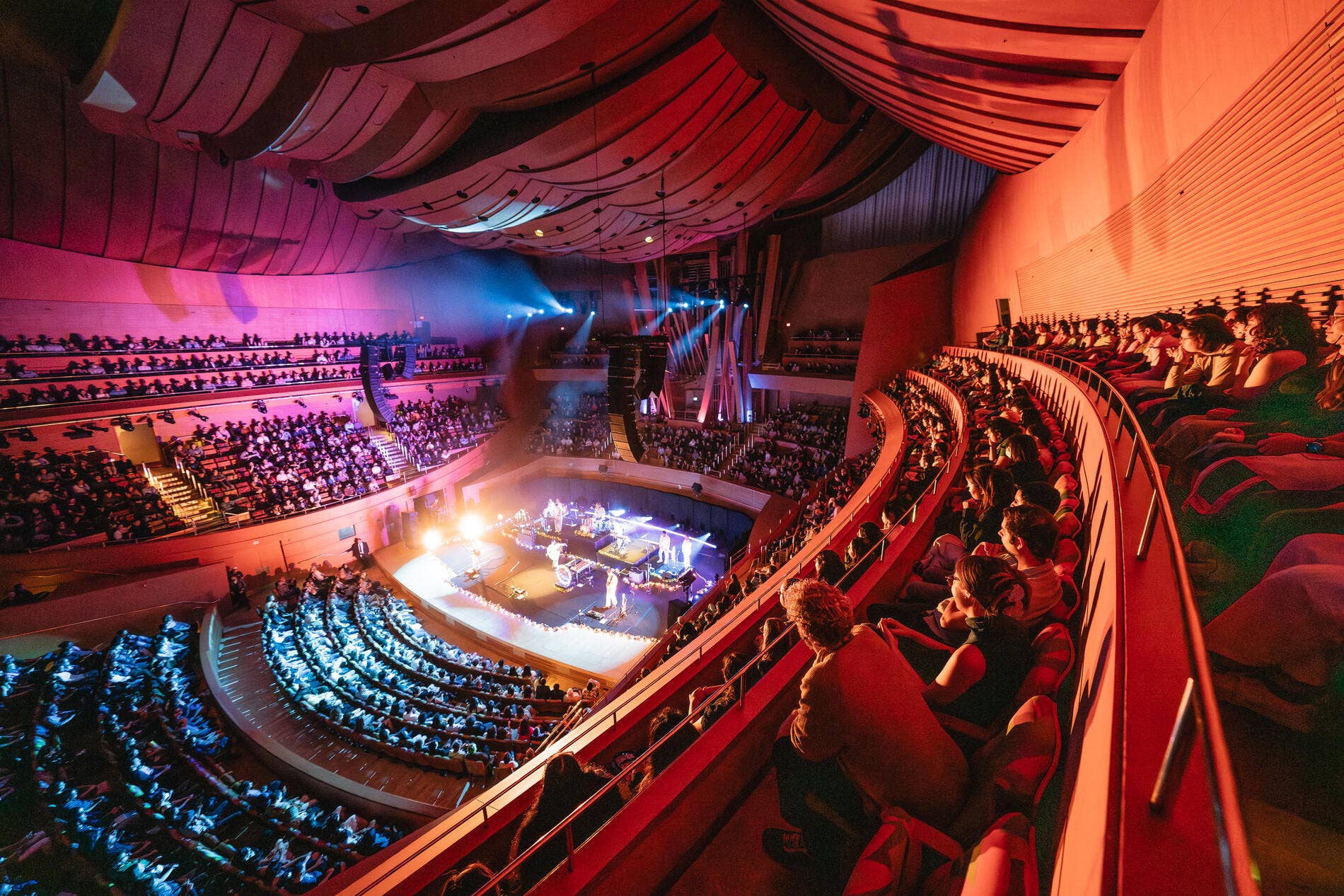Concert hall interior with curved red draped ceiling, spotlighted stage, and audience seated in balconies at night.