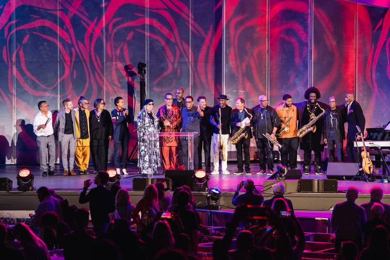 Several musicians stand on stage during a mid-performance speech.