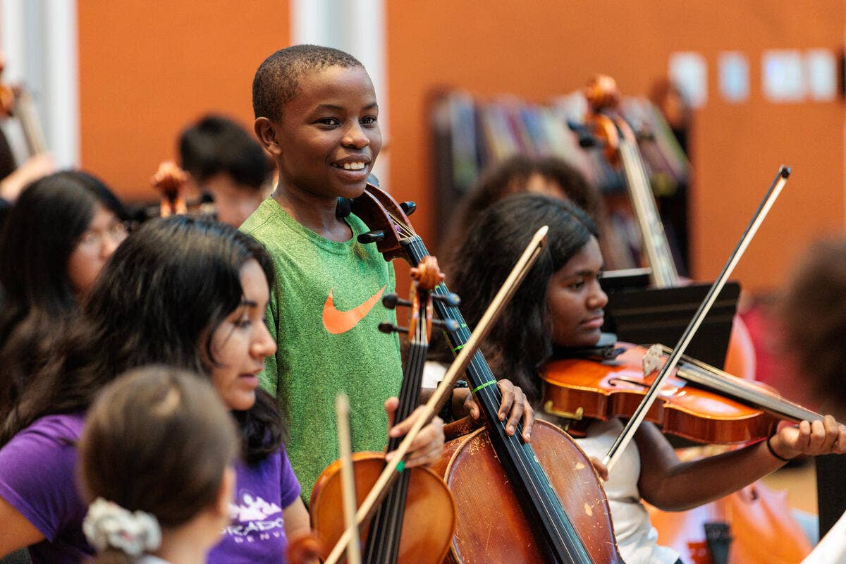 Un alumno sonriente, con una camiseta verde, toca el violín junto a sus compañeros durante un ensayo de la orquesta del colegio.