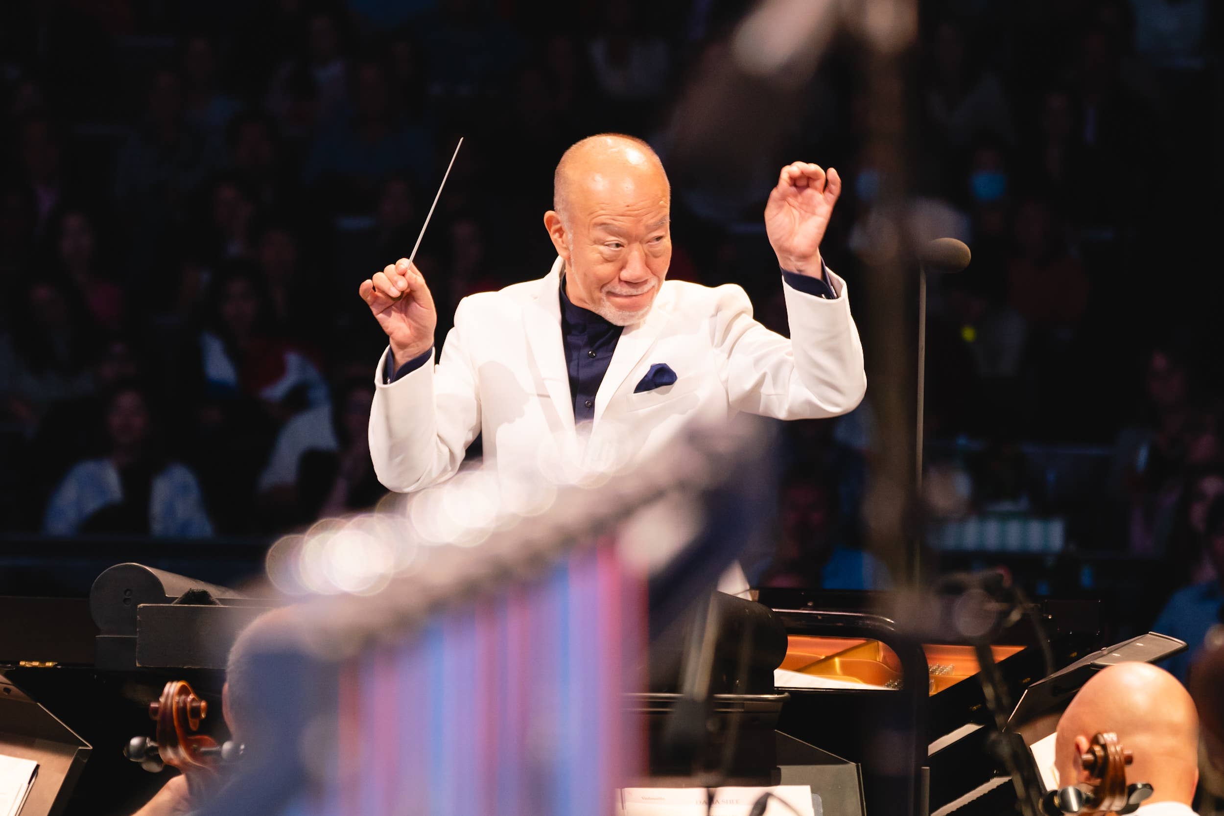 Smiling bald conductor in a white jacket raises a baton and gestures on a dark concert stage with orchestra musicians