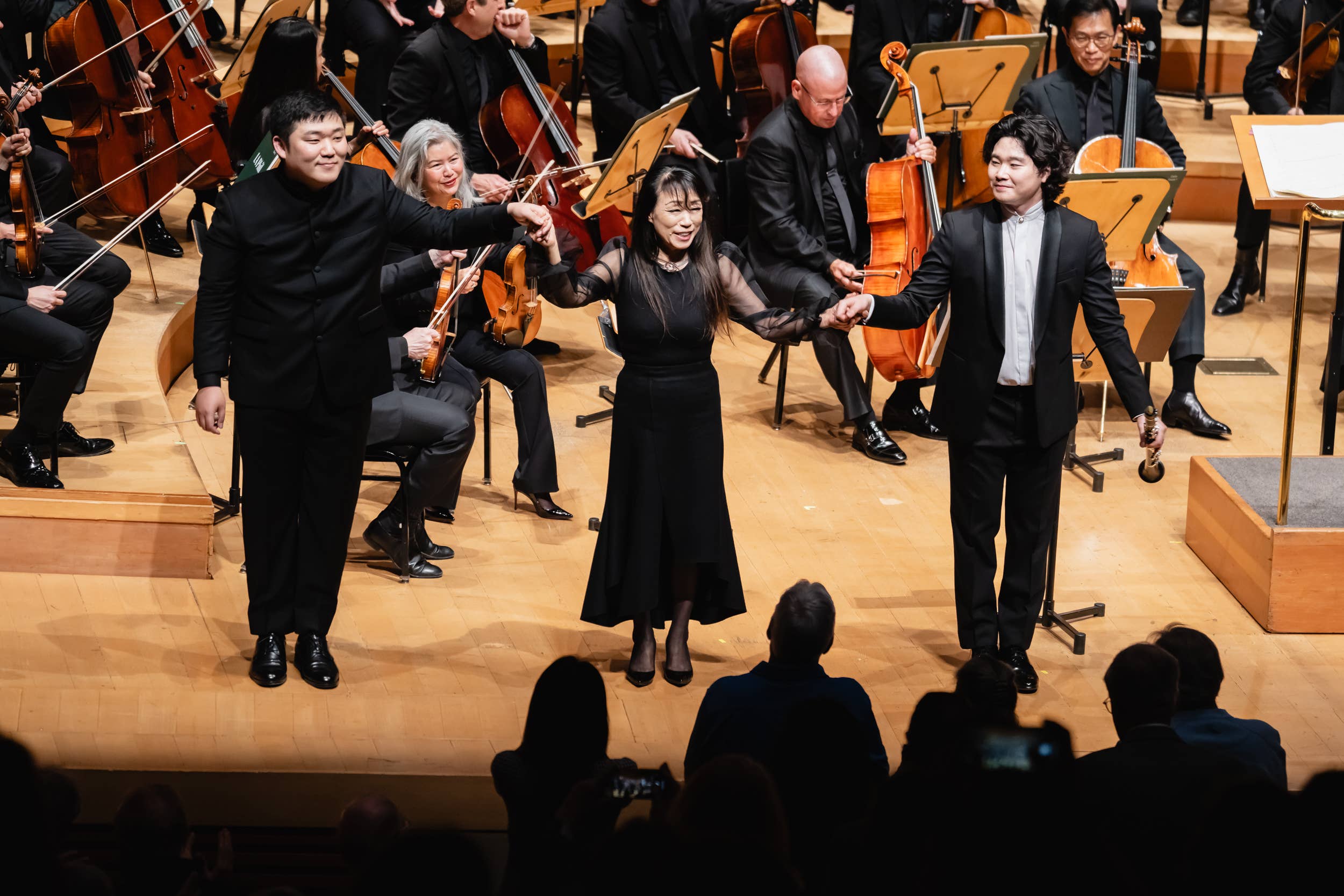 String quartet bowing onstage with cellists and violins, conductor nearby, audience silhouettes in foreground