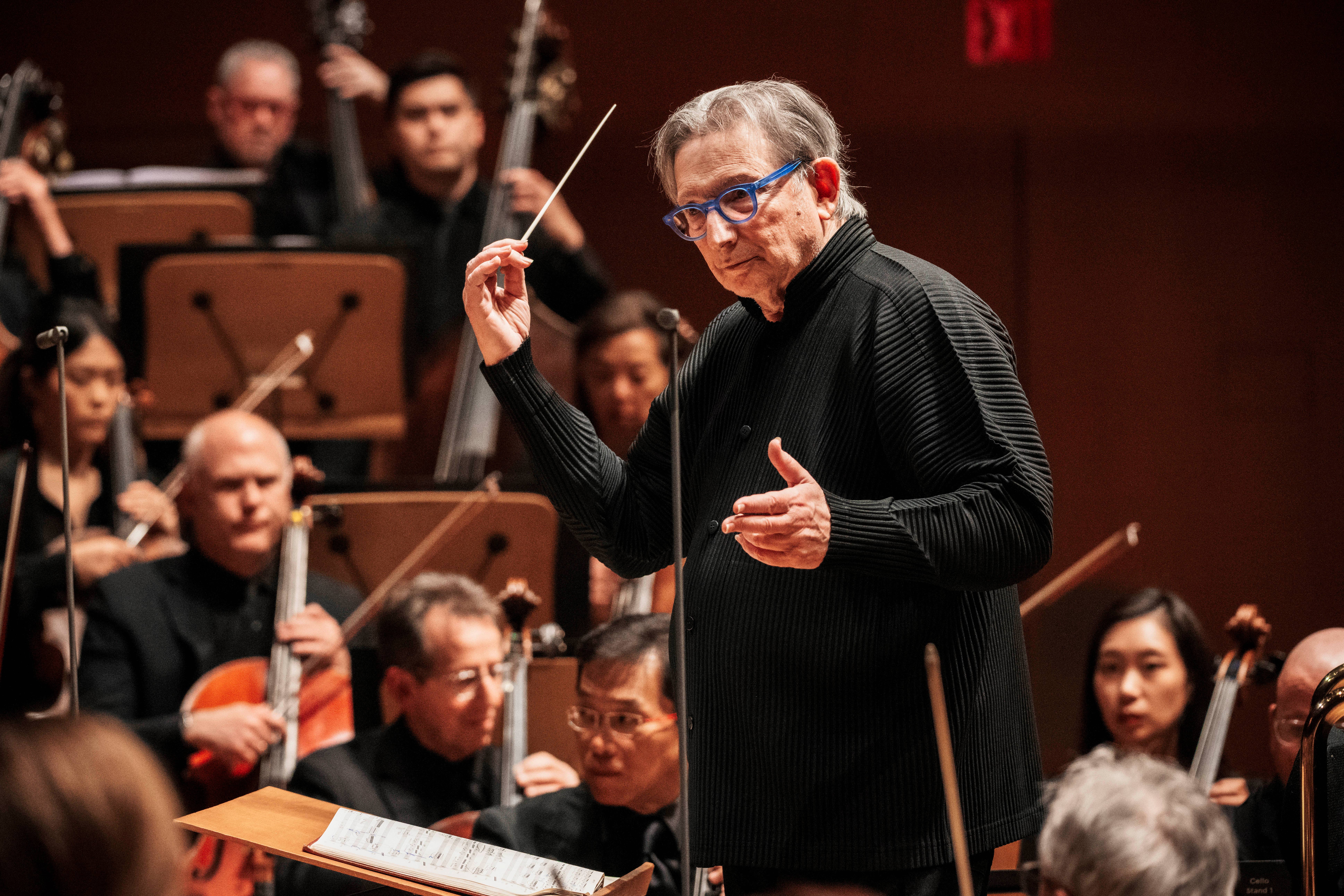 director de orquesta de edad avanzada, director de orquesta gafas azules y batuta en la mano, dirige una orquesta cuyos músicos están sentados detrás de él.