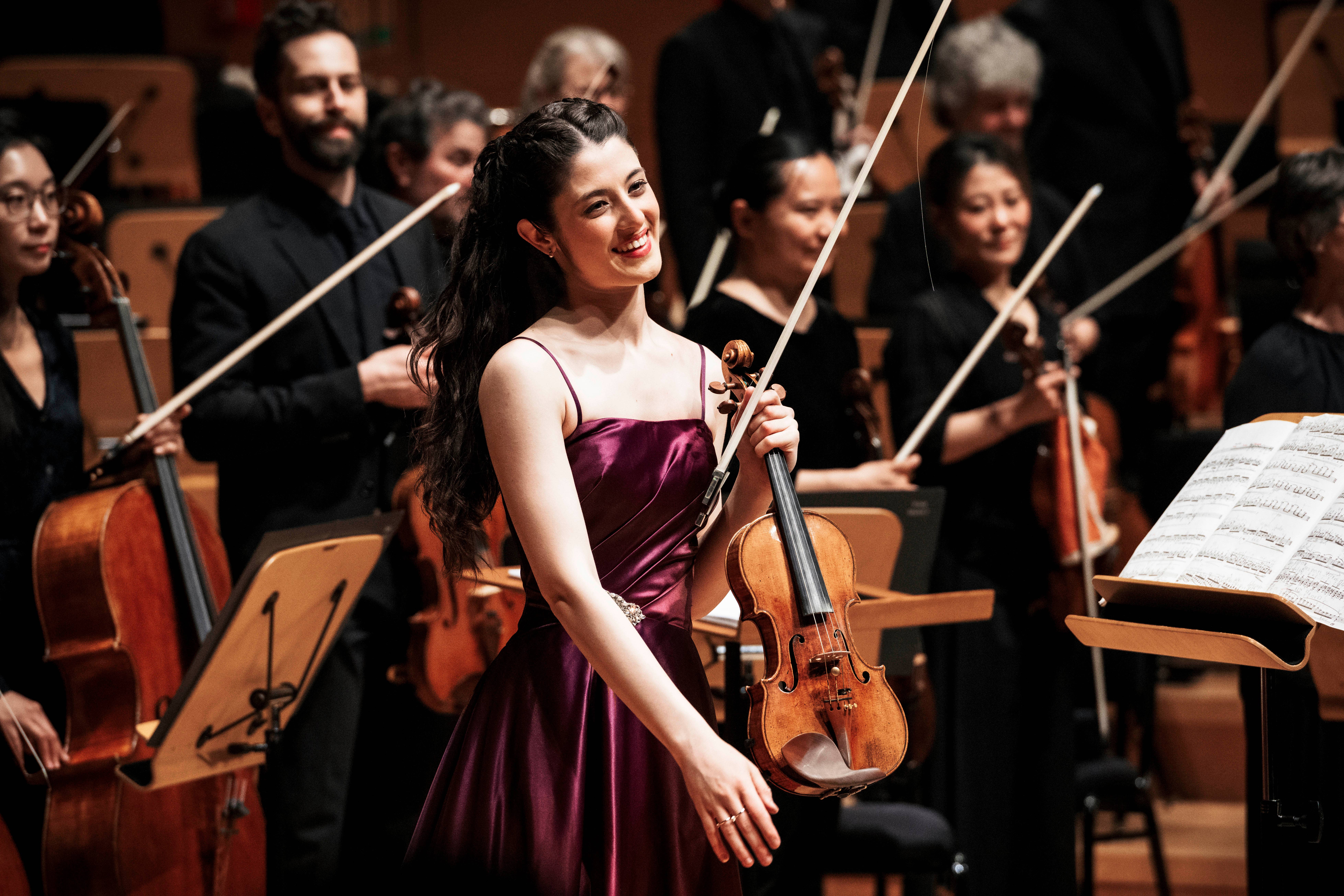 Smiling violinist in a purple dress performing on stage with an orchestra behind her, bows and sheet music visible