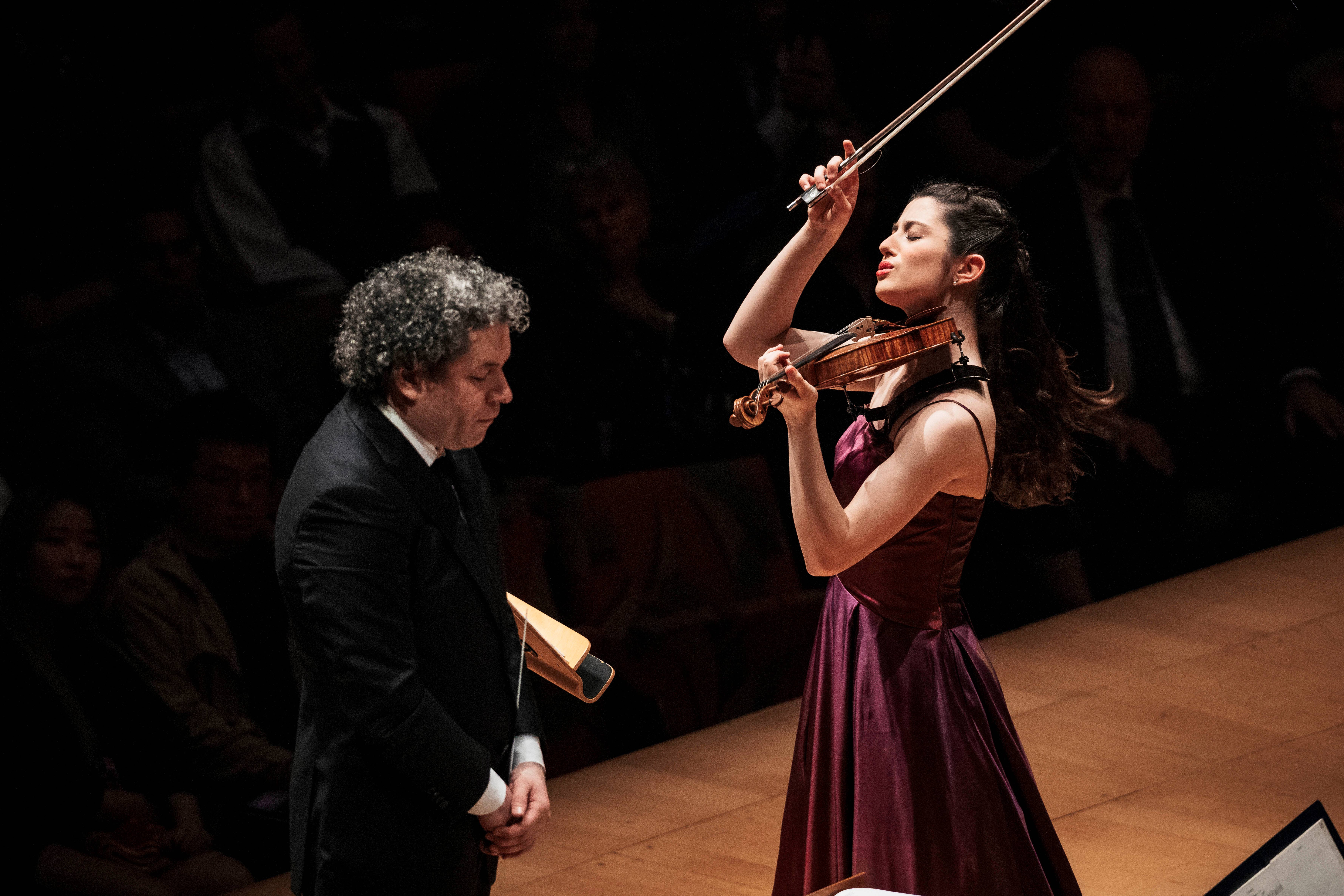 Female violinist in a burgundy dress plays with bow raised while conductor listens in a dark concert hall