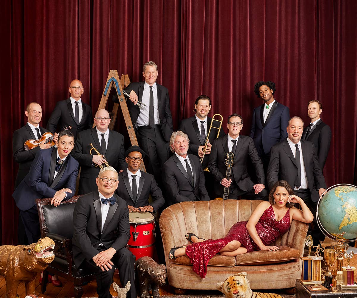 Members of Pink Martini are dressed in formal attire in front of a red, velvet curtain. 