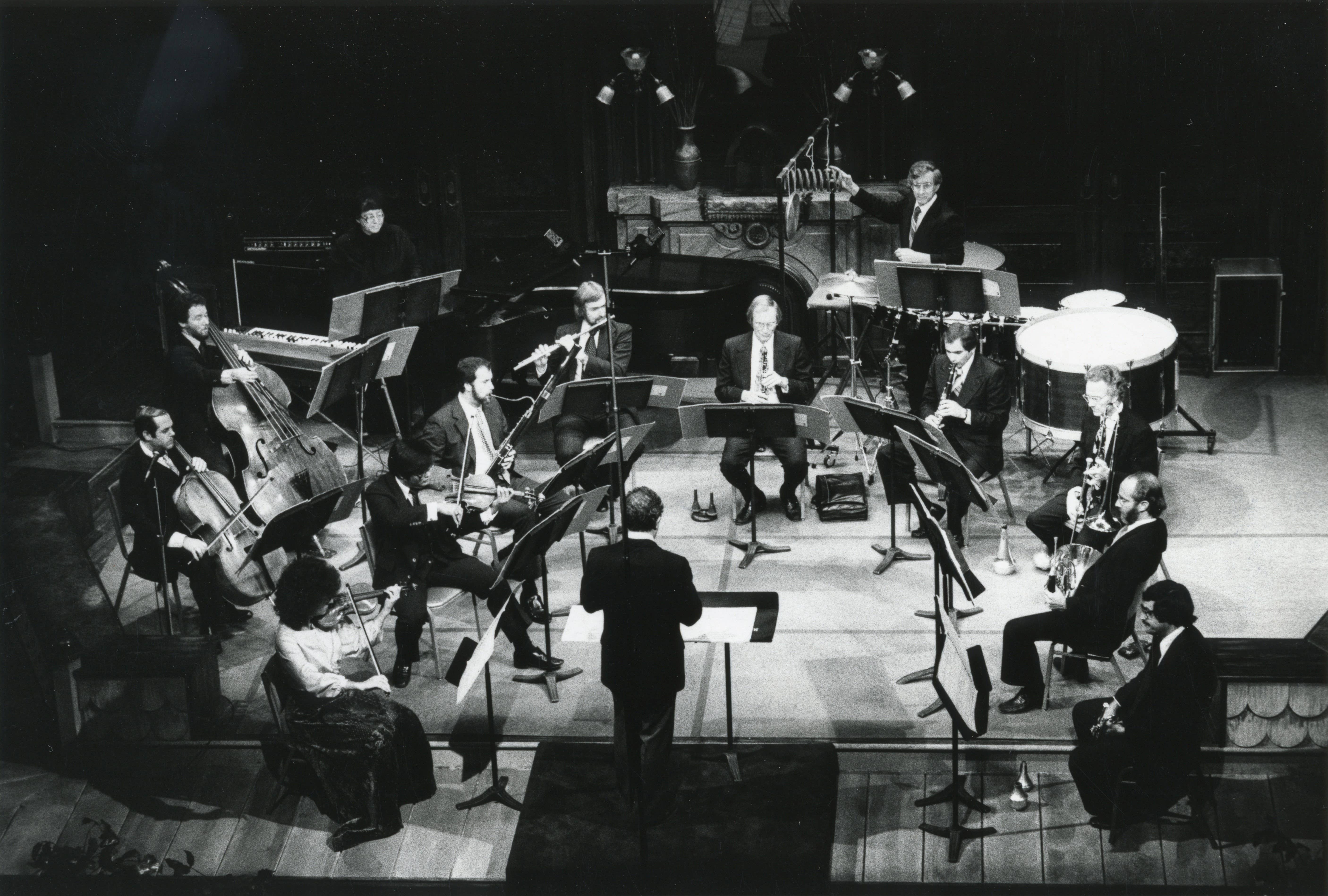Fotografía en blanco y negro de una orquesta sinfónica actuando en el escenario con director de orquesta, los metales, las cuerdas y los timbales.