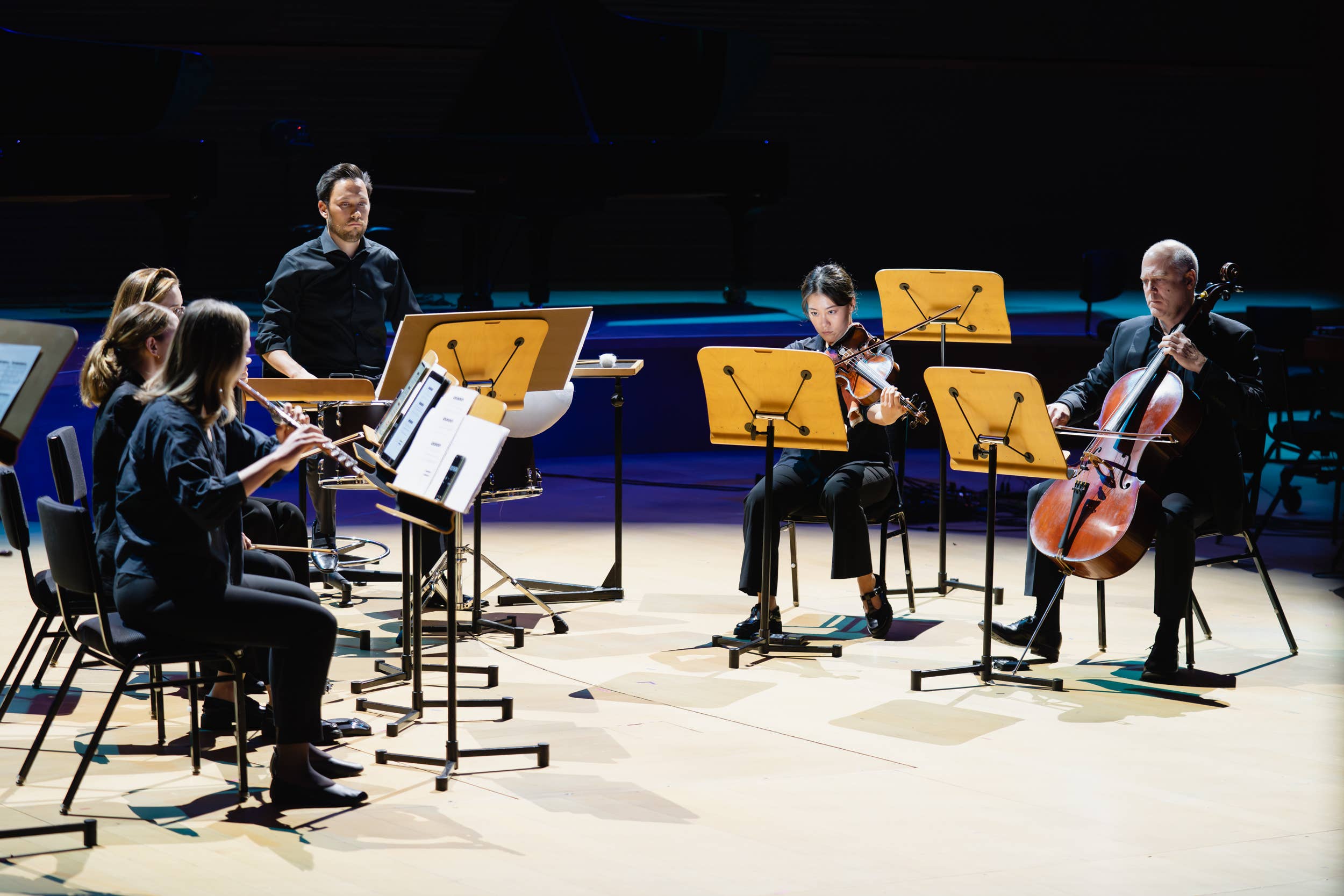 Ensayo de una orquesta en una sala de conciertos, con músicos que sostienen un violín y una flauta entre atriles y un director de orquesta.
