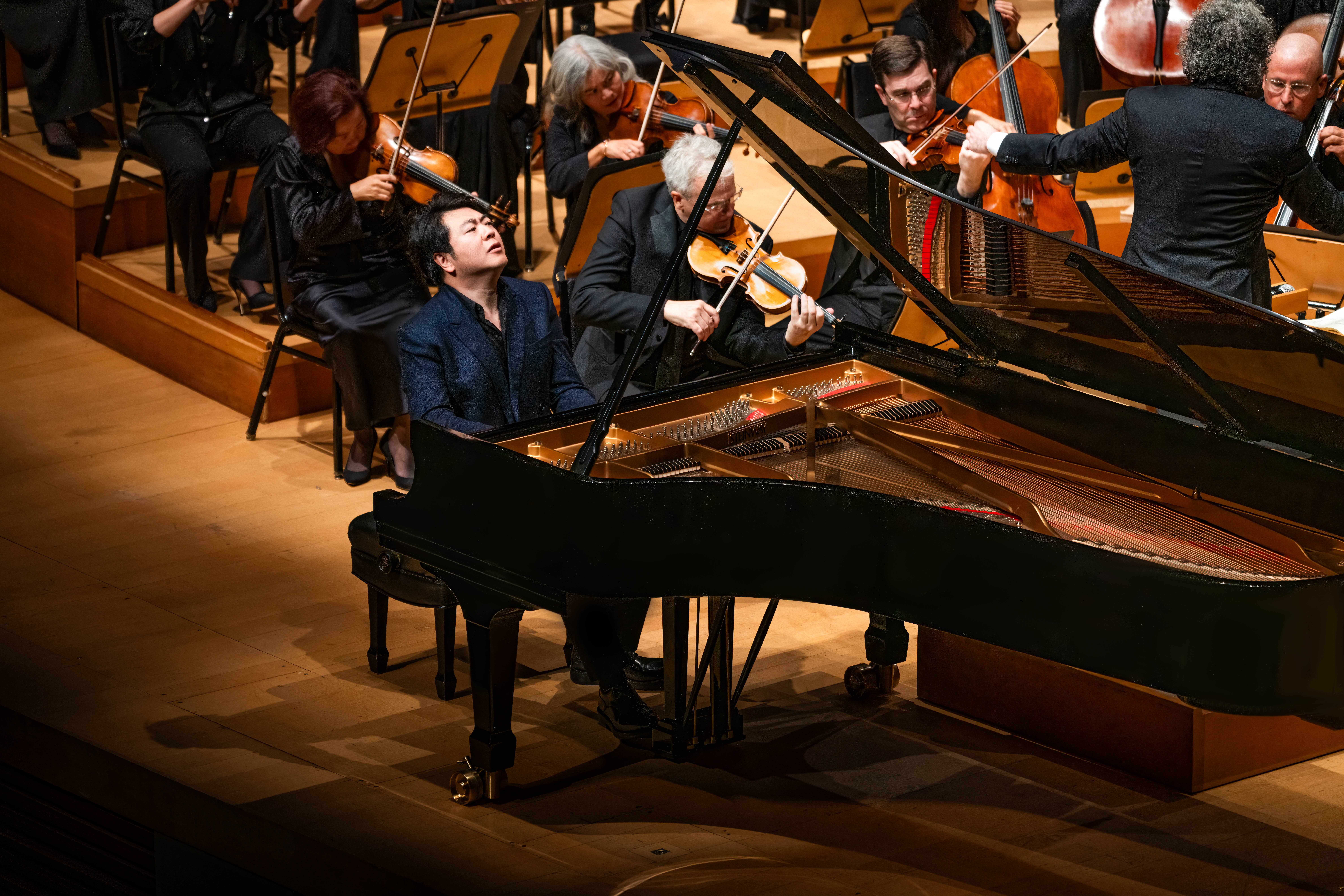 Los músicos de la orquesta tocan junto a una mujer que está al piano de cola piano el escenario de un concierto.