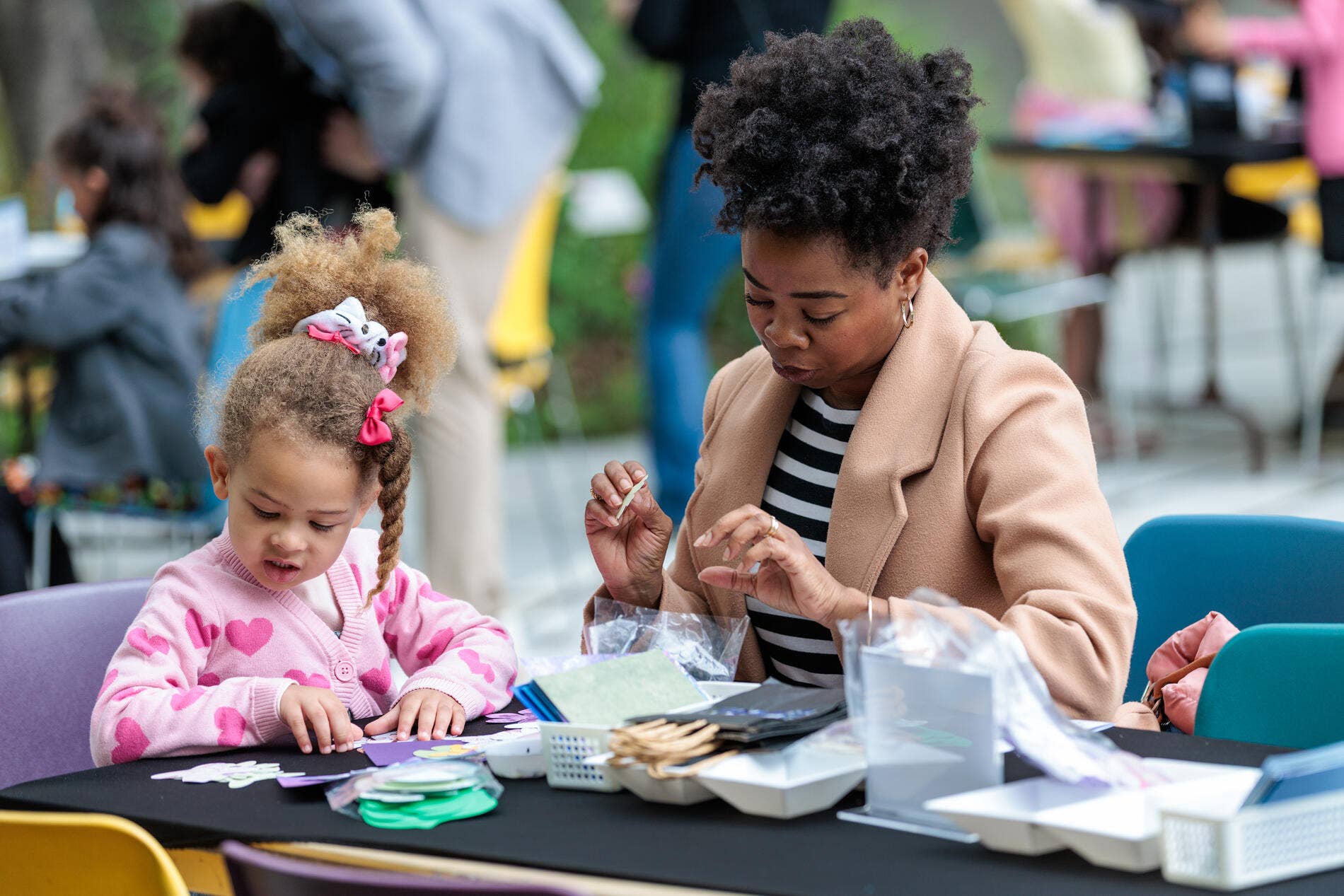 Two young children craft at a table while a woman assists, focused on paper and art supplies outdoors.