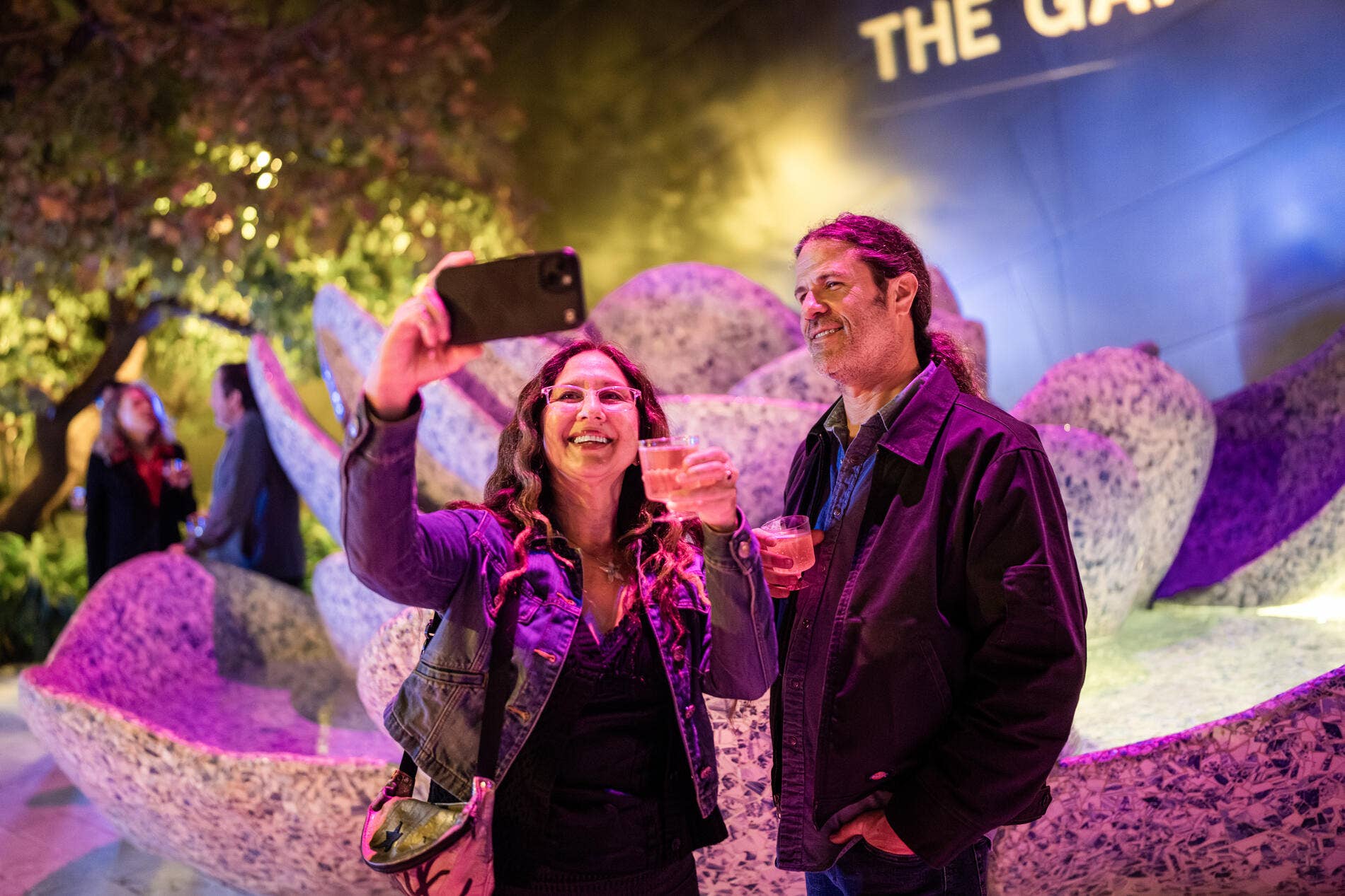 Smiling couple at an outdoor event, taking a selfie while holding drinks amid colorful stage lighting