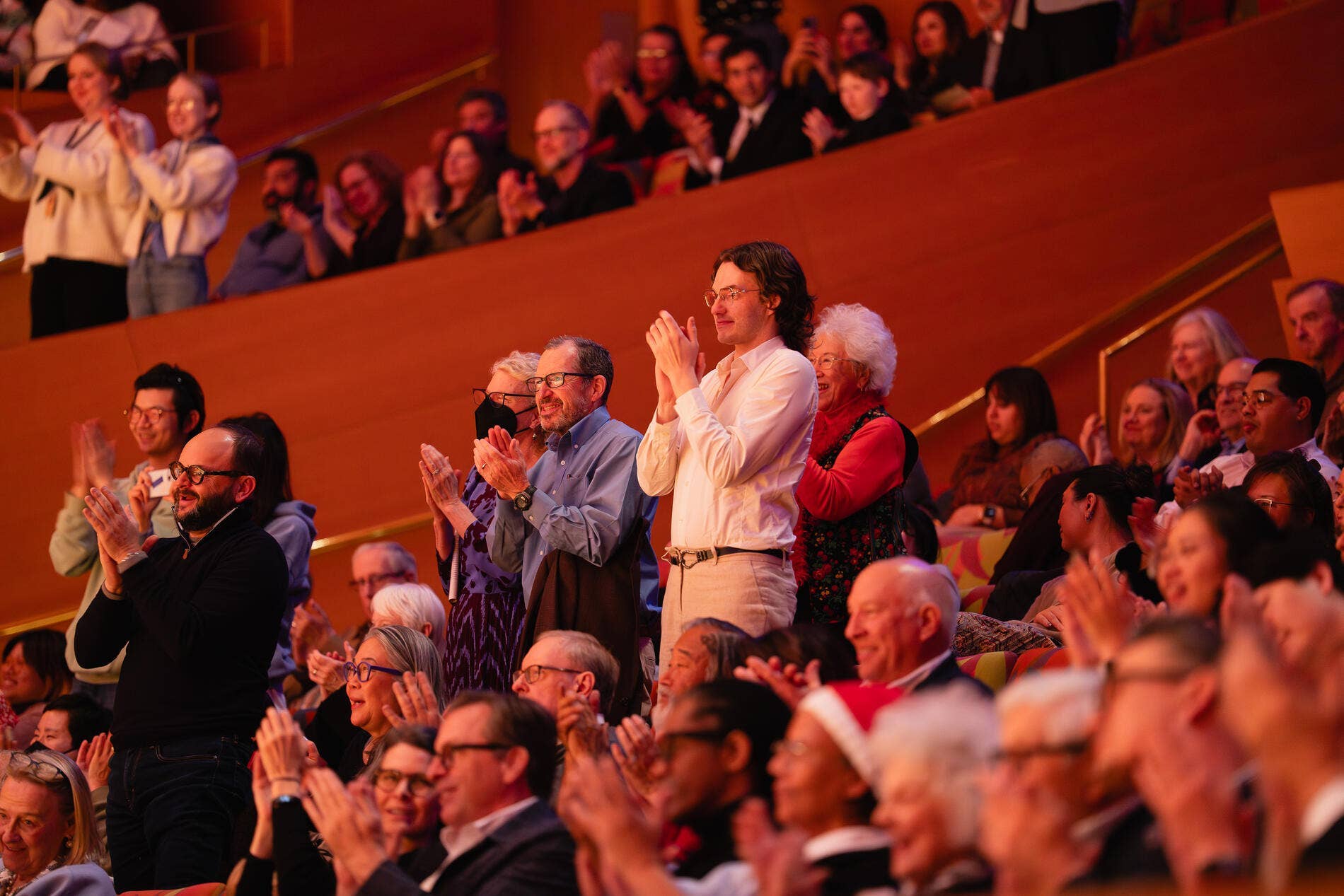 Audience applauds in a theater auditorium, with people standing and clapping under warm red lighting.