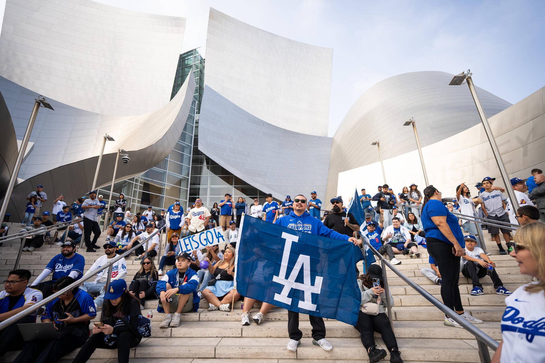 Aficionados de los Dodgers de Los Ángeles en un estadio, animando y ondeando una pancarta azul frente a unos modernos edificios blancos.