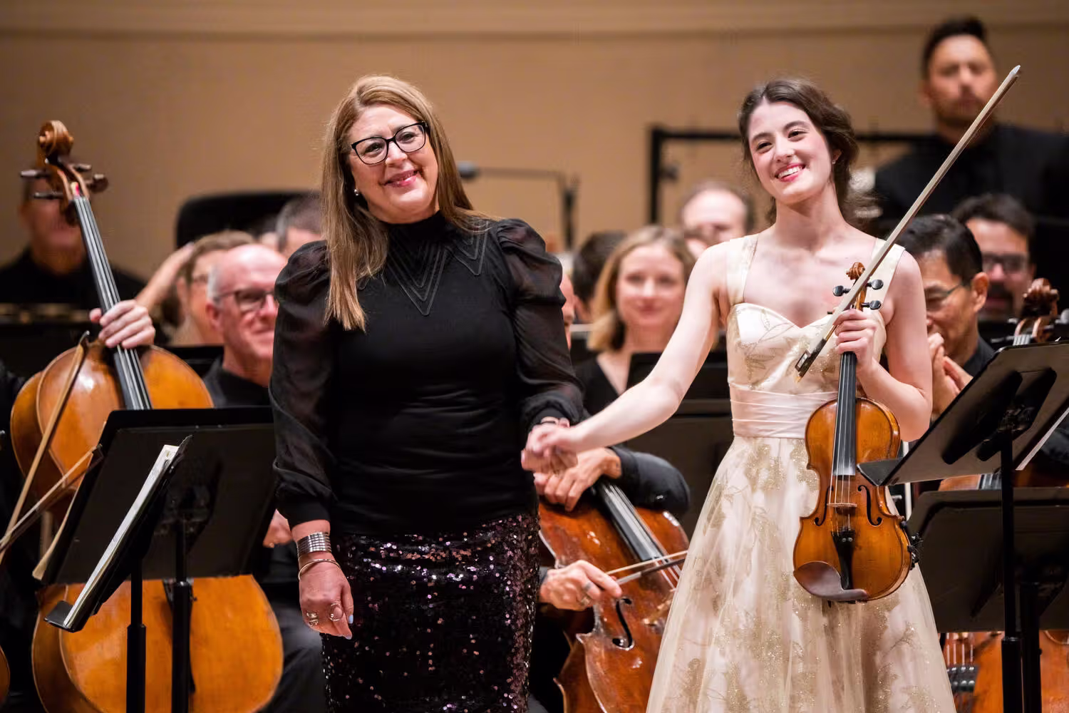 Two musicians in a concert hall, one conductor with baton and another violinist playing amid an orchestra.