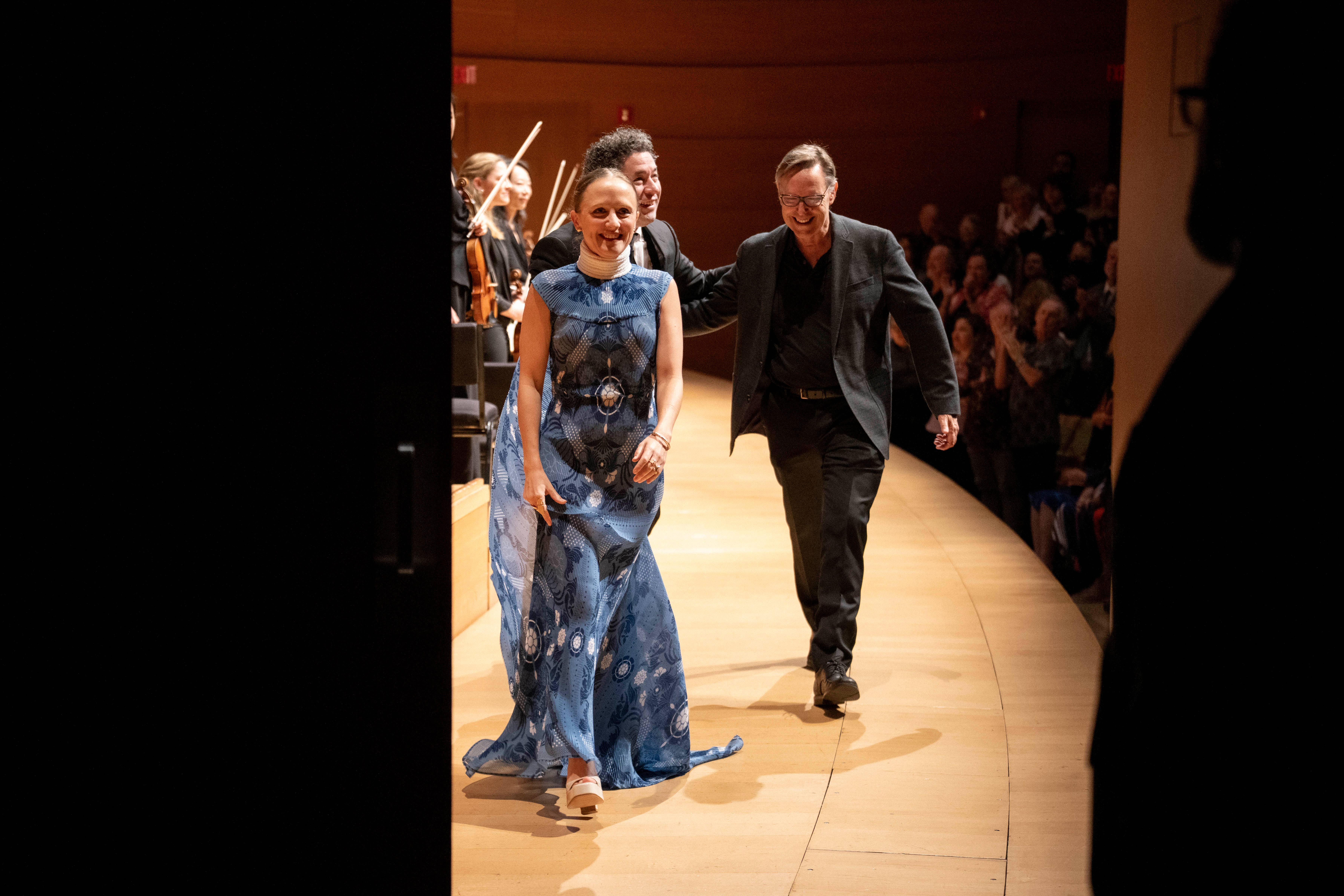Woman in blue gown and scarf walks arm-in-arm with man in dark suit during runway event, smiling.