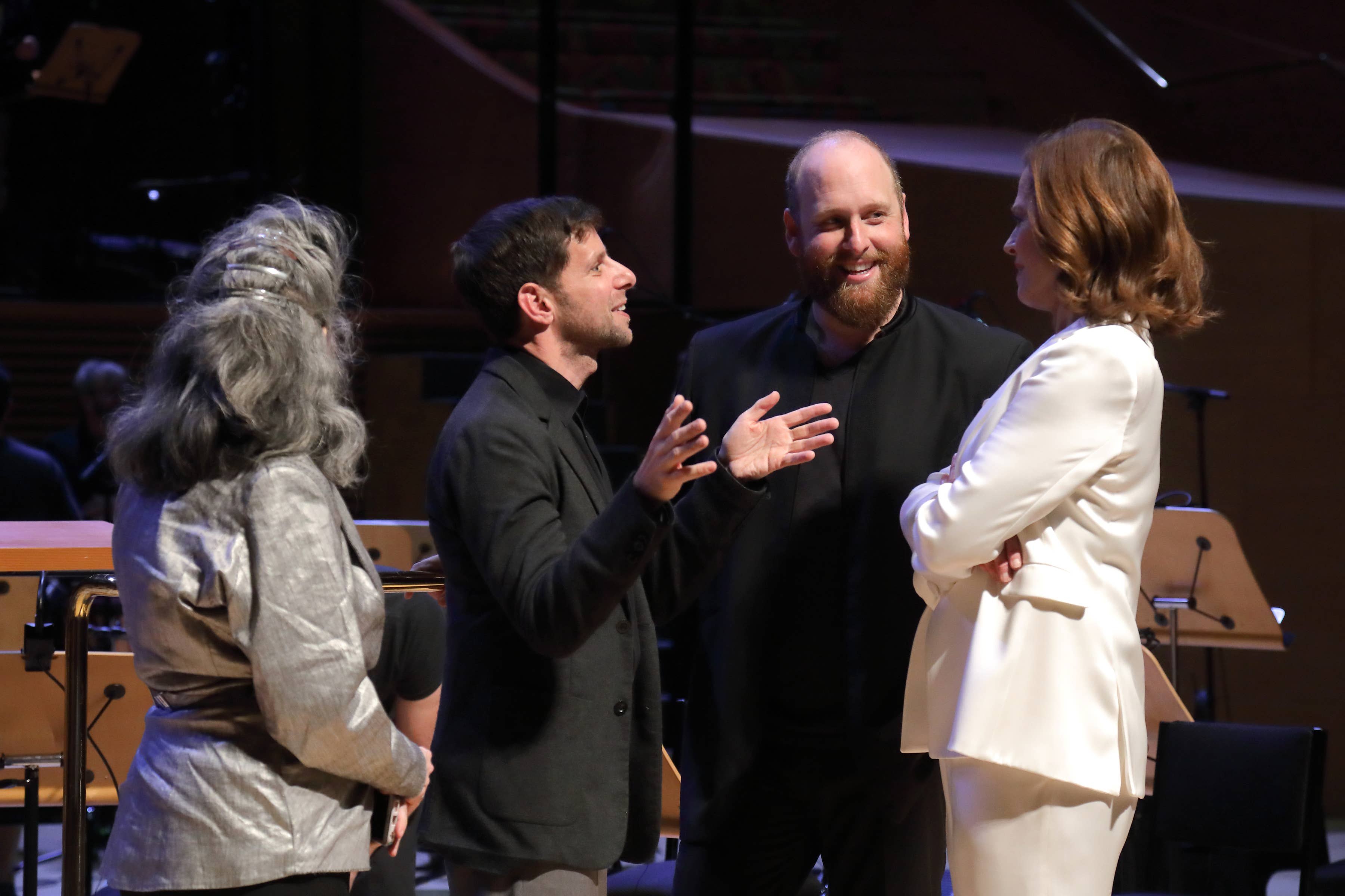 Cuatro artistas en el escenario sonríen y gesticulan mientras un hombre con chaqueta oscura habla.