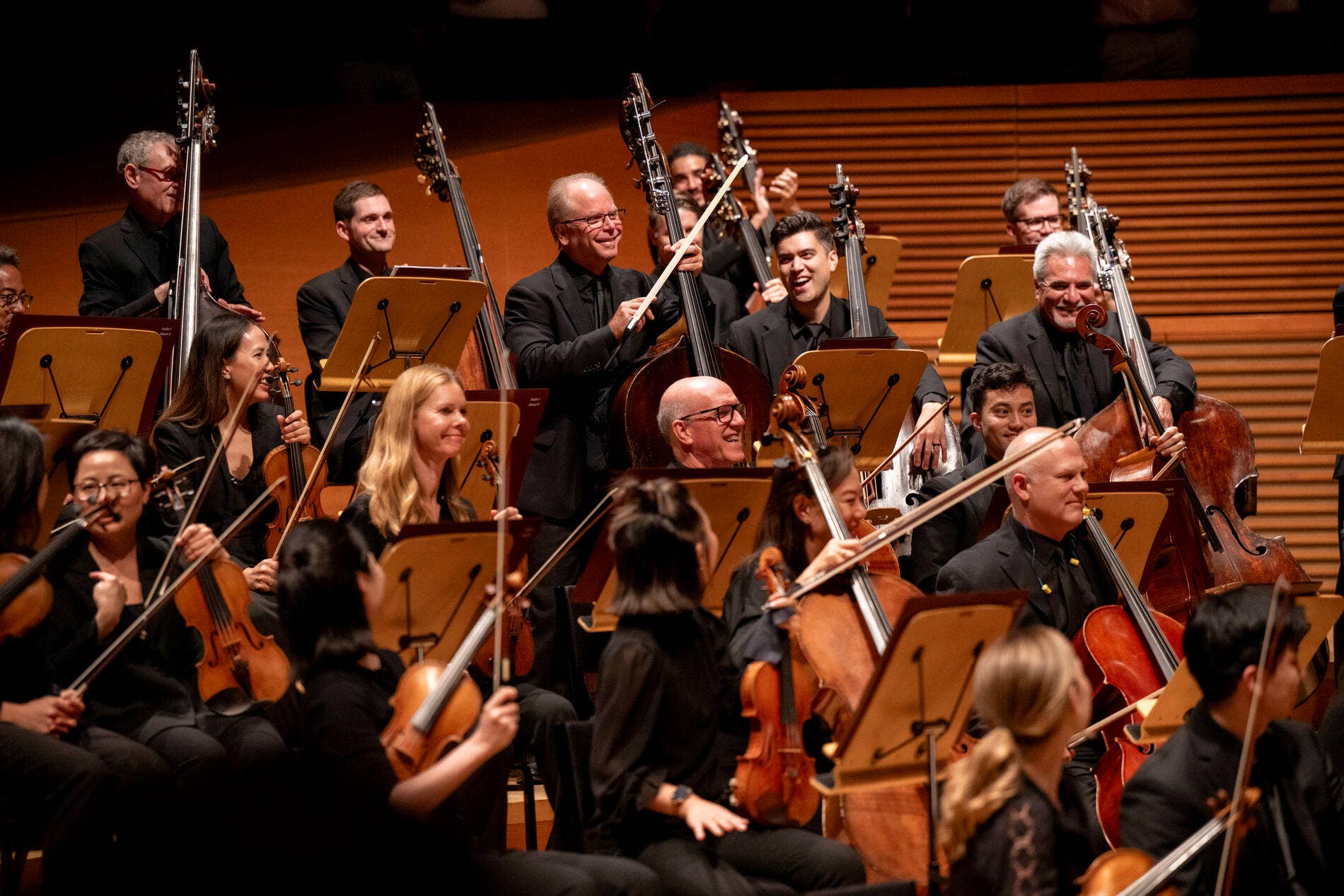 Orchestra musicians in black attire perform, smiling with upright basses, violins, and clarinets on stage in a concert hall
