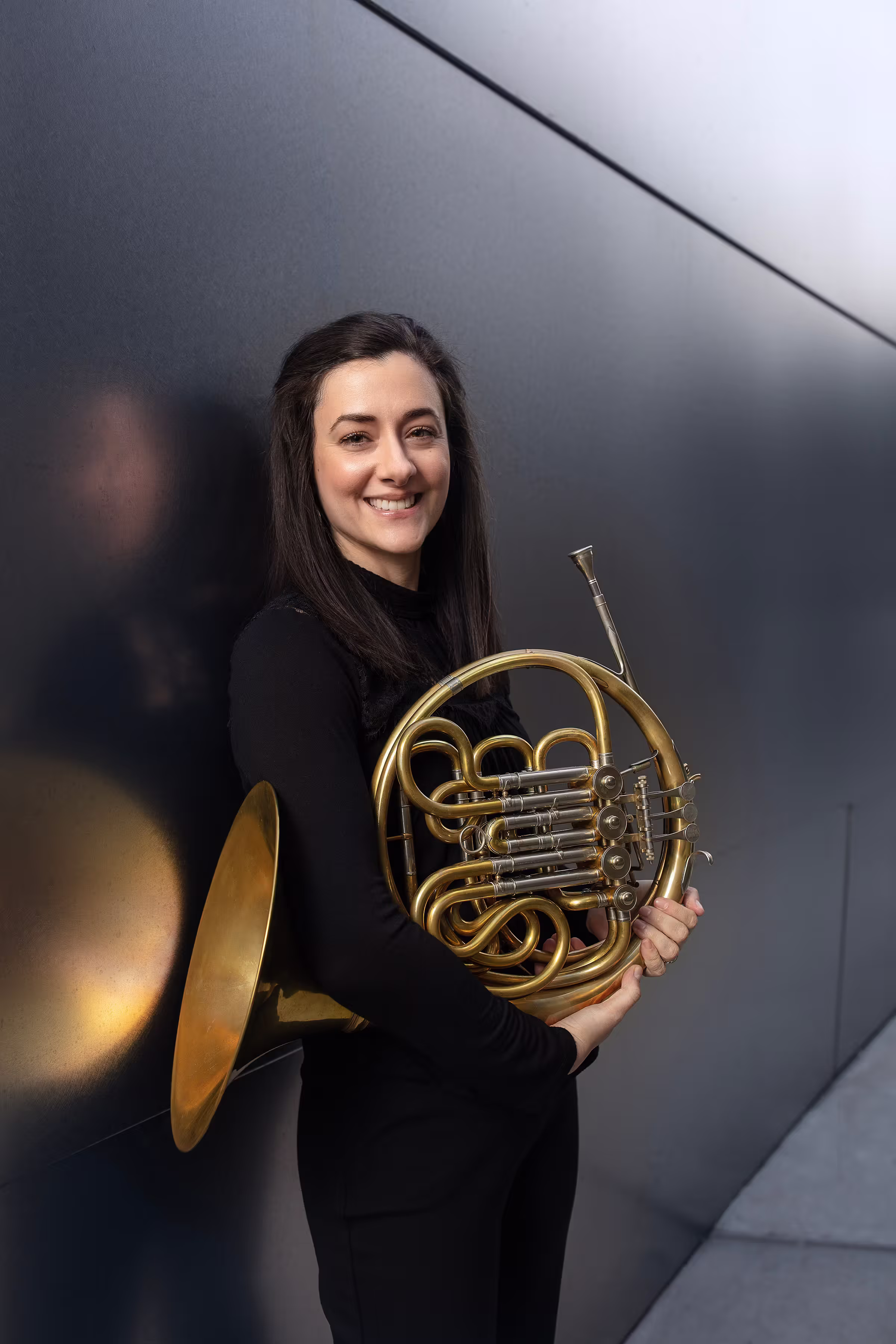 Smiling woman holding a brass French horn in a studio portrait against a gray backdrop