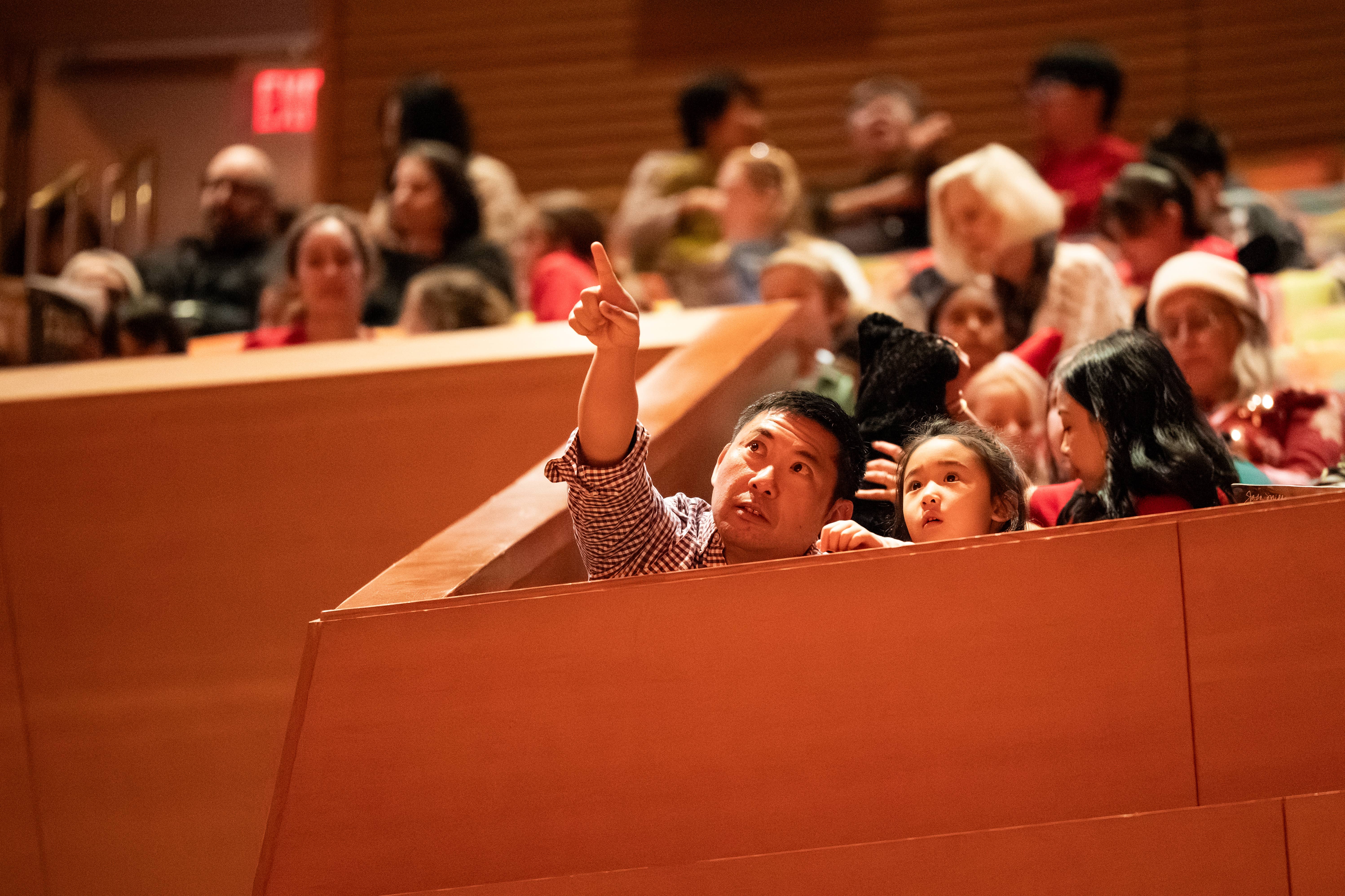 Audience in a theater balcony; a man raises his hand as a child and others watch.
