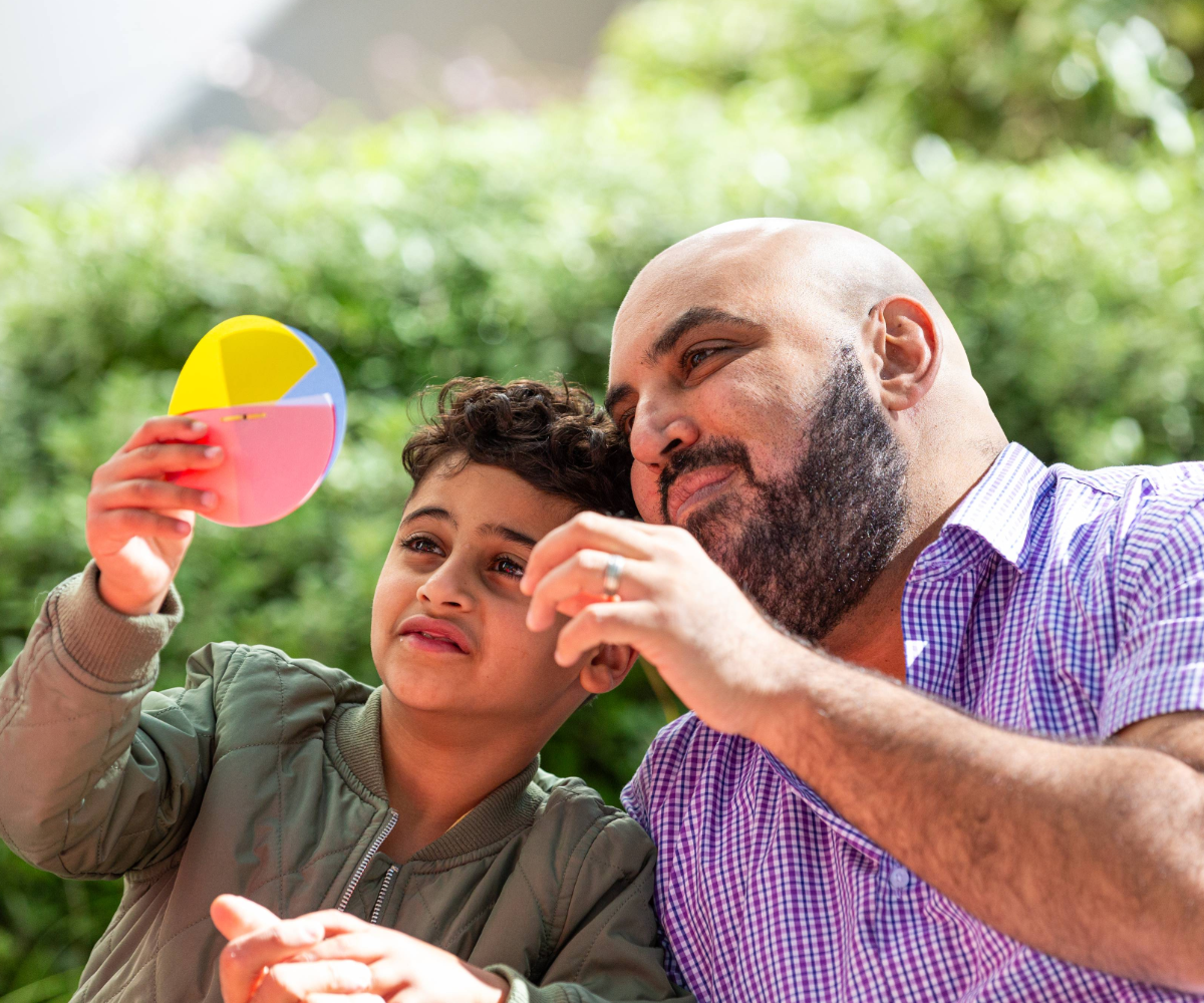 Man and boy play outdoors, with boy holding a colorful hand mirror toward his father in sunny greenery.
