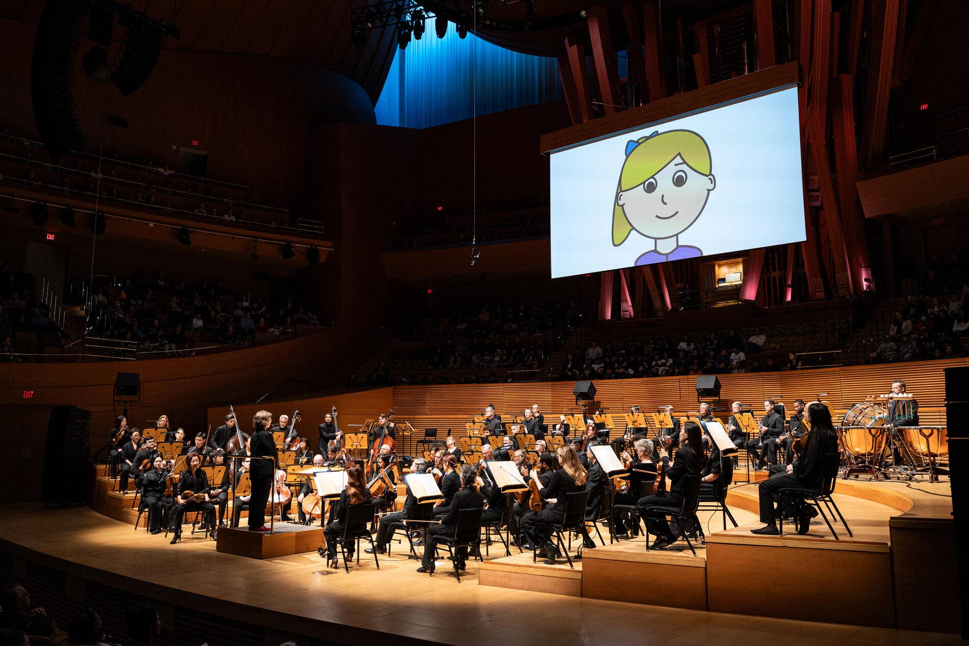 Orchestra performance in a concert hall with a cartoon girl on a large screen above musicians and instruments.