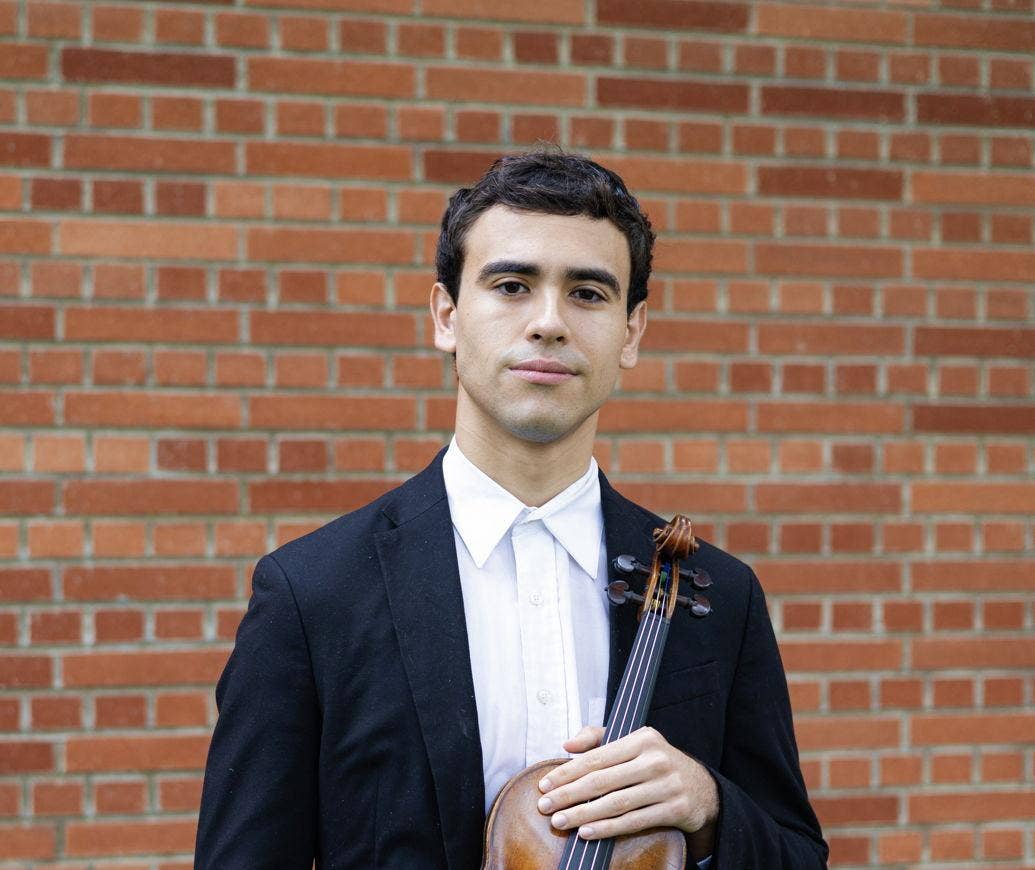 Un hombre con traje negro que sostiene un violín, con una pared de ladrillos al fondo, mirando a la cámara.