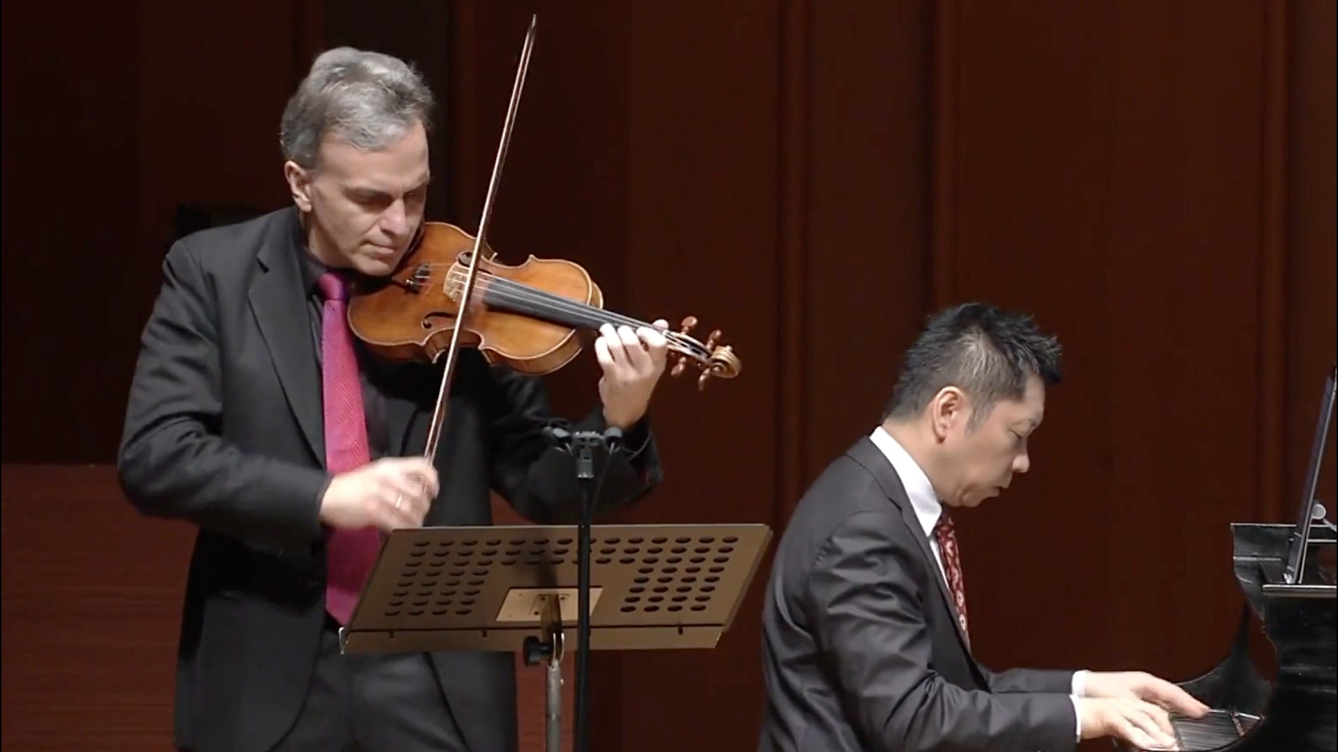 Violinist in suit playing on stage as a pianist sits nearby in a concert hall.