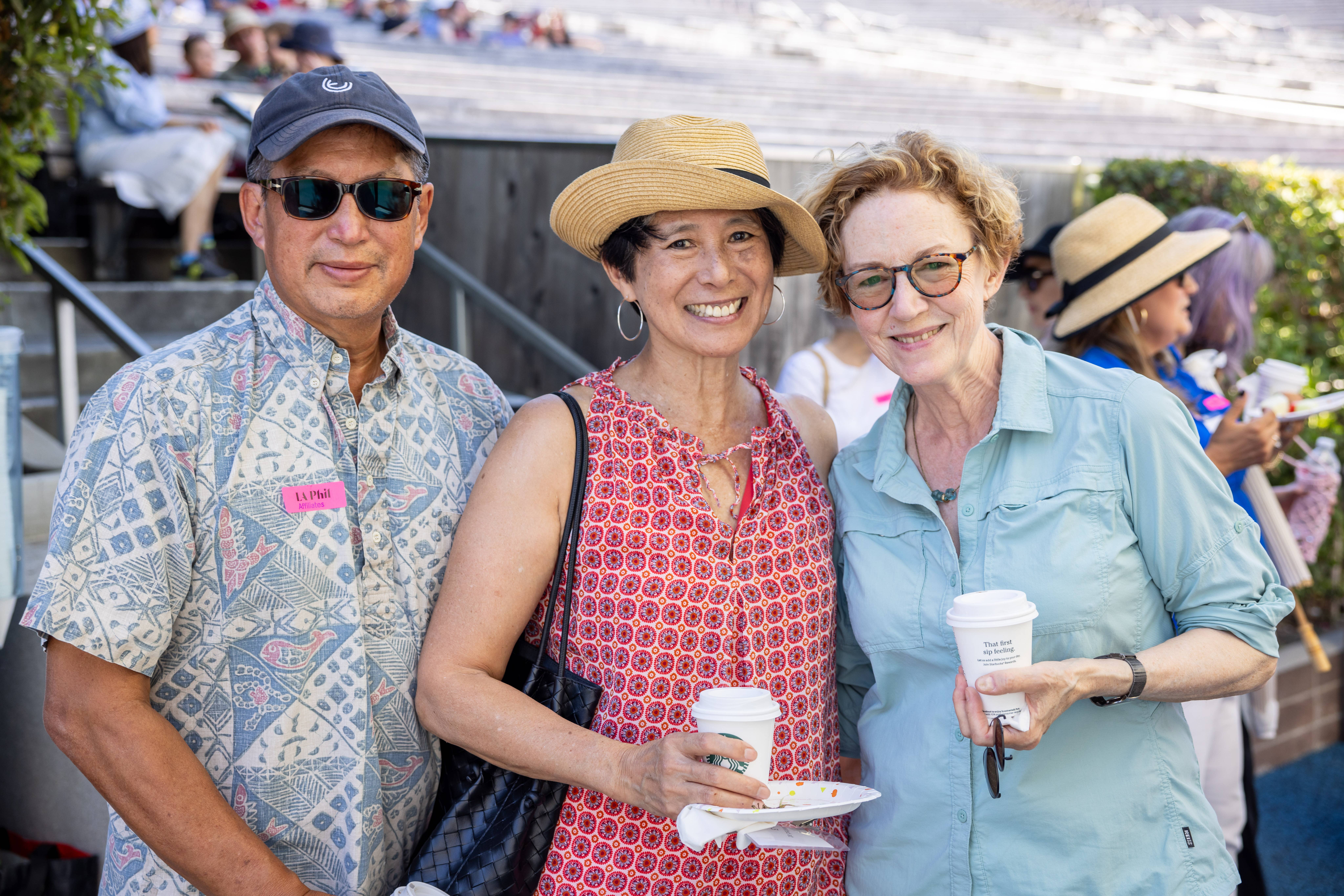 Tres adultos sonriendo al aire libre en un evento, con tazas de café en la mano, gorras y gafas de sol puestas