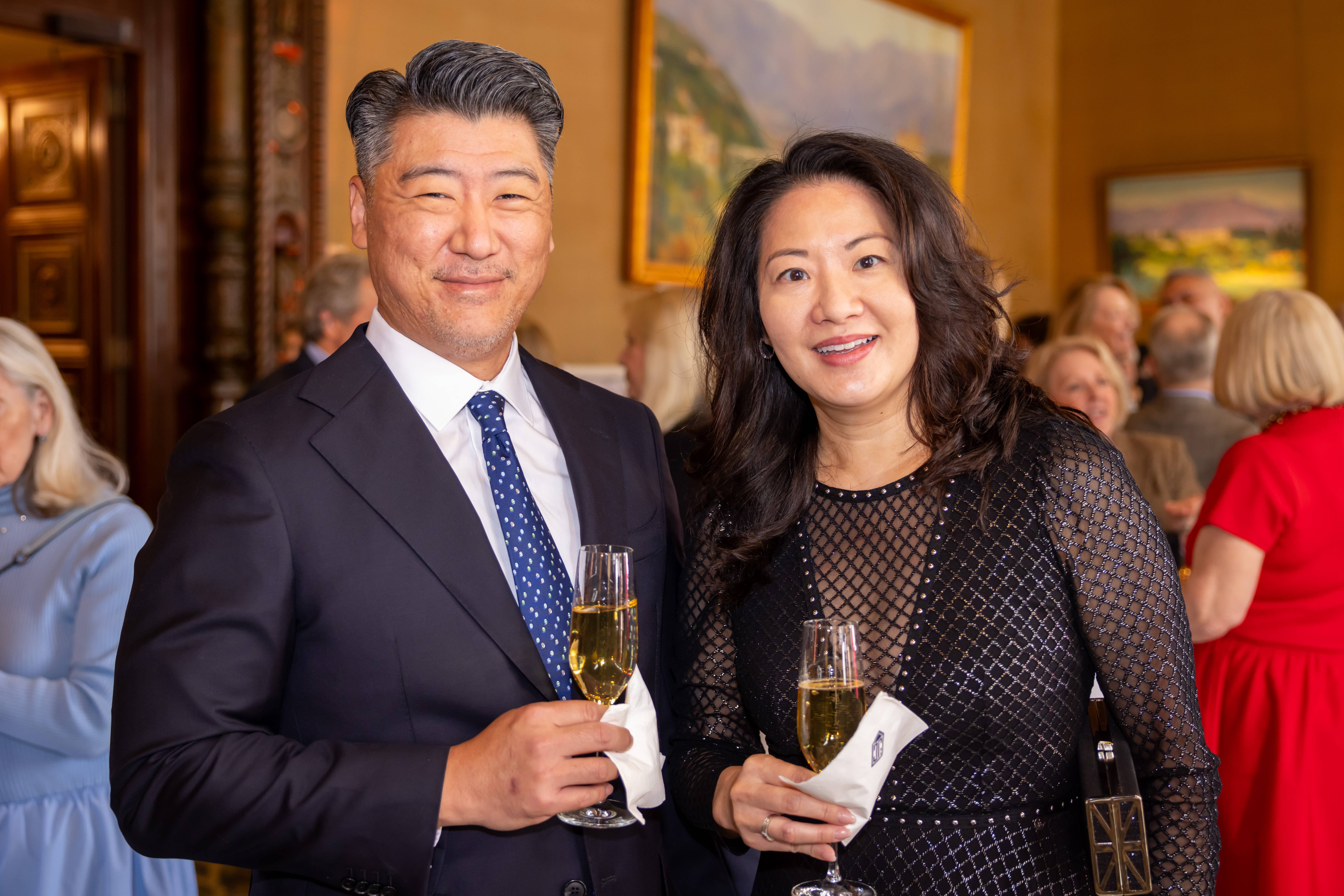 Pareja sonriente, vestida de gala, sosteniendo copas de champán en una recepción en un salón, con los invitados al fondo.