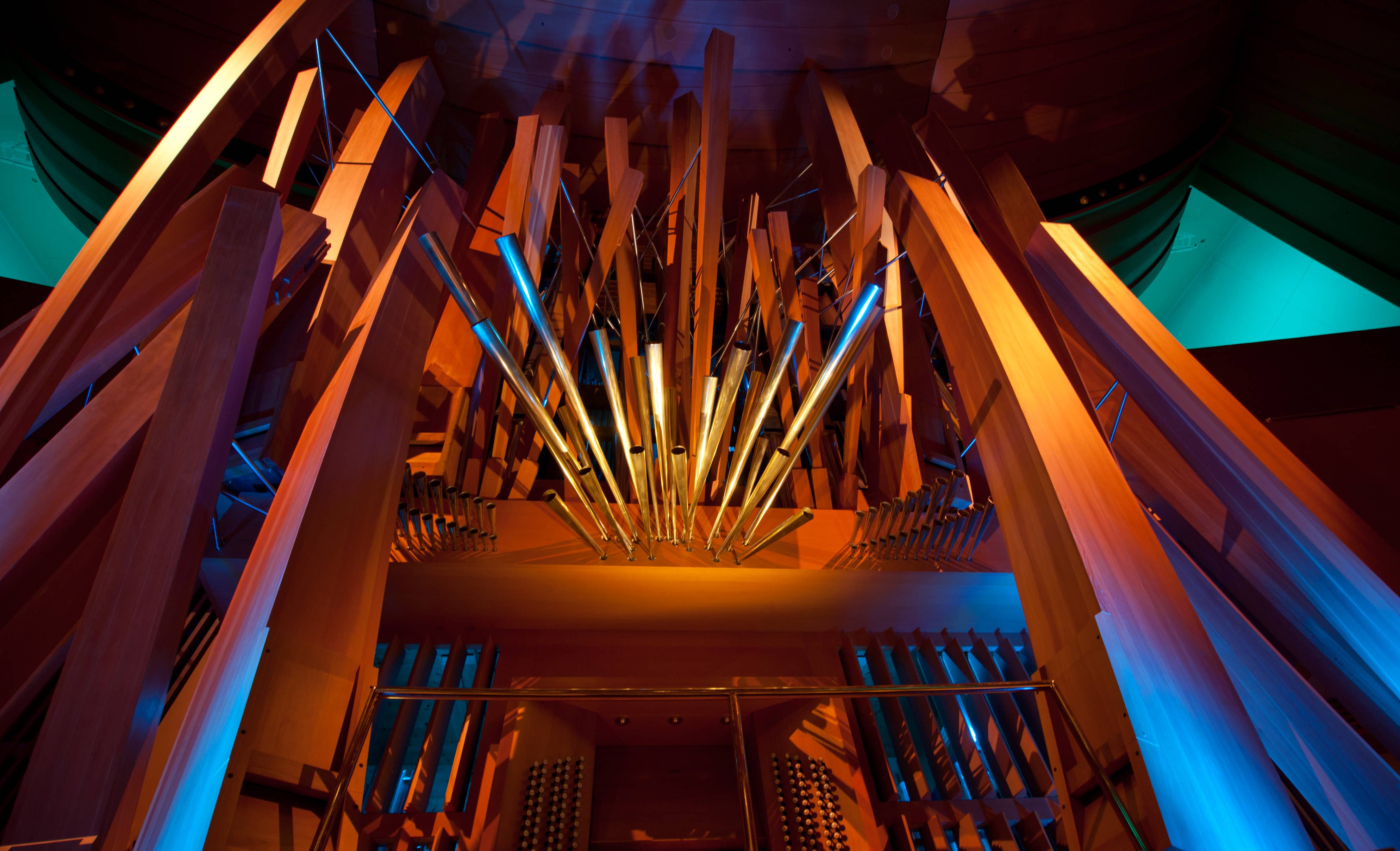 Colorful pipe organ interior with rows of brass pipes and wooden beams under blue-orange lighting.