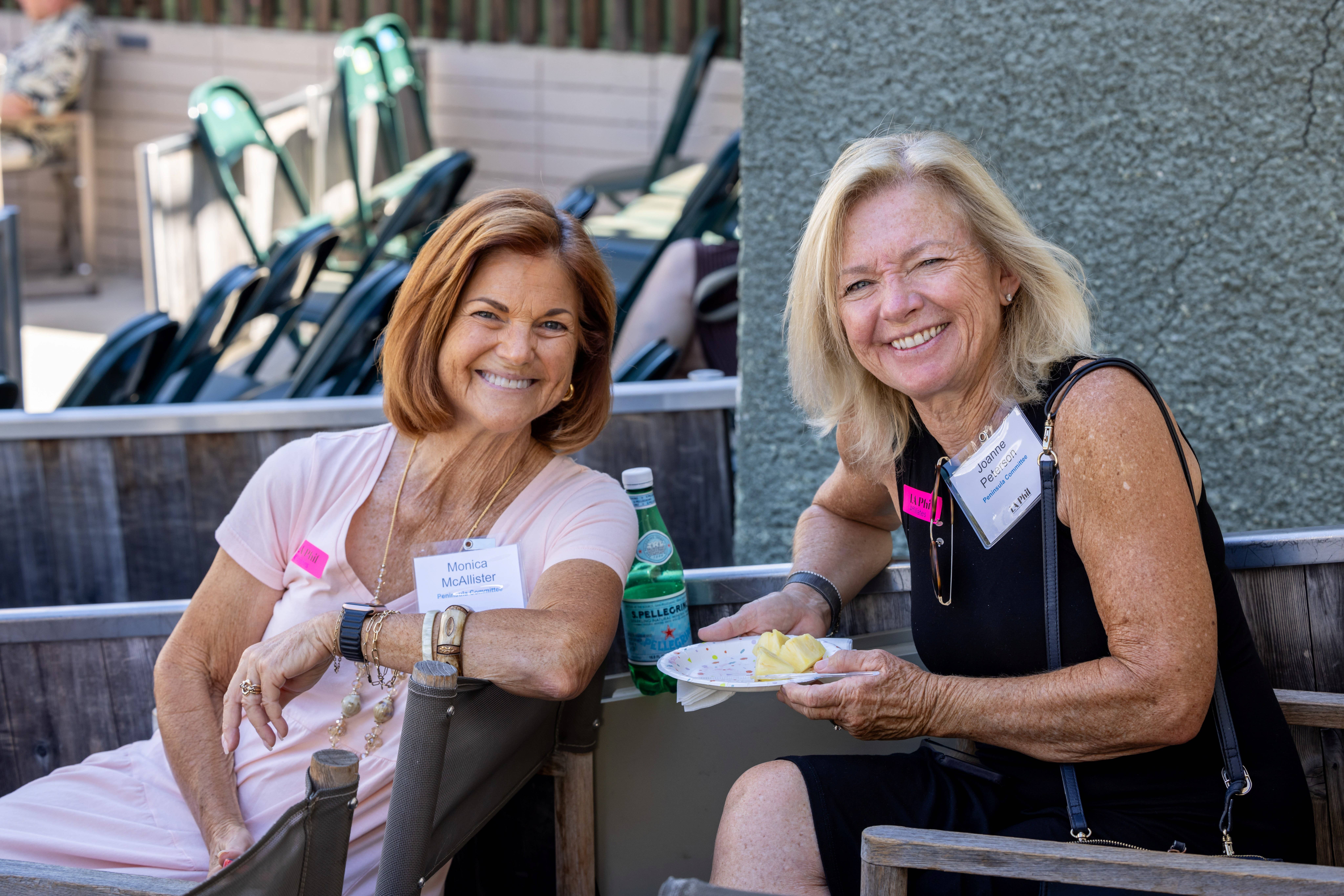 Dos mujeres sonrientes sentadas al aire libre en una mesa de un evento, con insignias, bebidas y sillas detrás de ellas