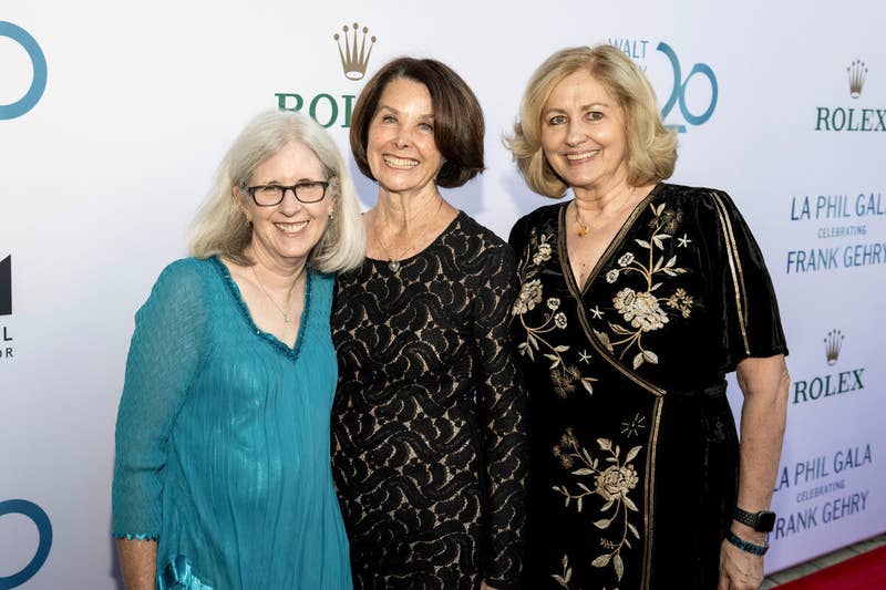 Tres mujeres posan en una gala de La Phil ante un telón de fondo con un Rolex y una obra de Frank Gehry.
