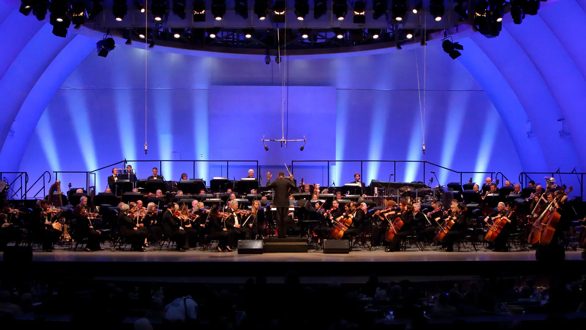 Large concert hall orchestra performing onstage under blue spotlights with musicians seated in rows.