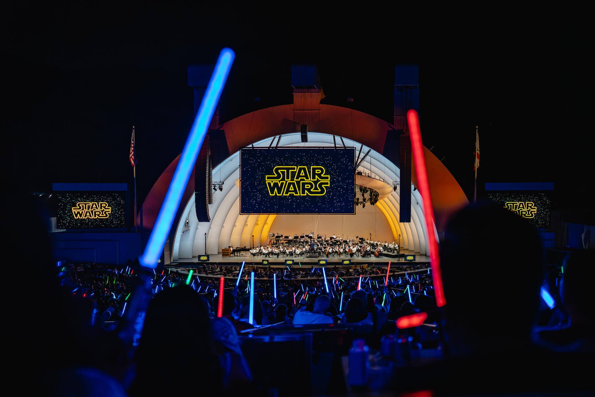 Escenario de un concierto de «Star Wars» con orquesta, sables láser azules y rojos que brillan y pantallas con el logotipo de «Star Wars».